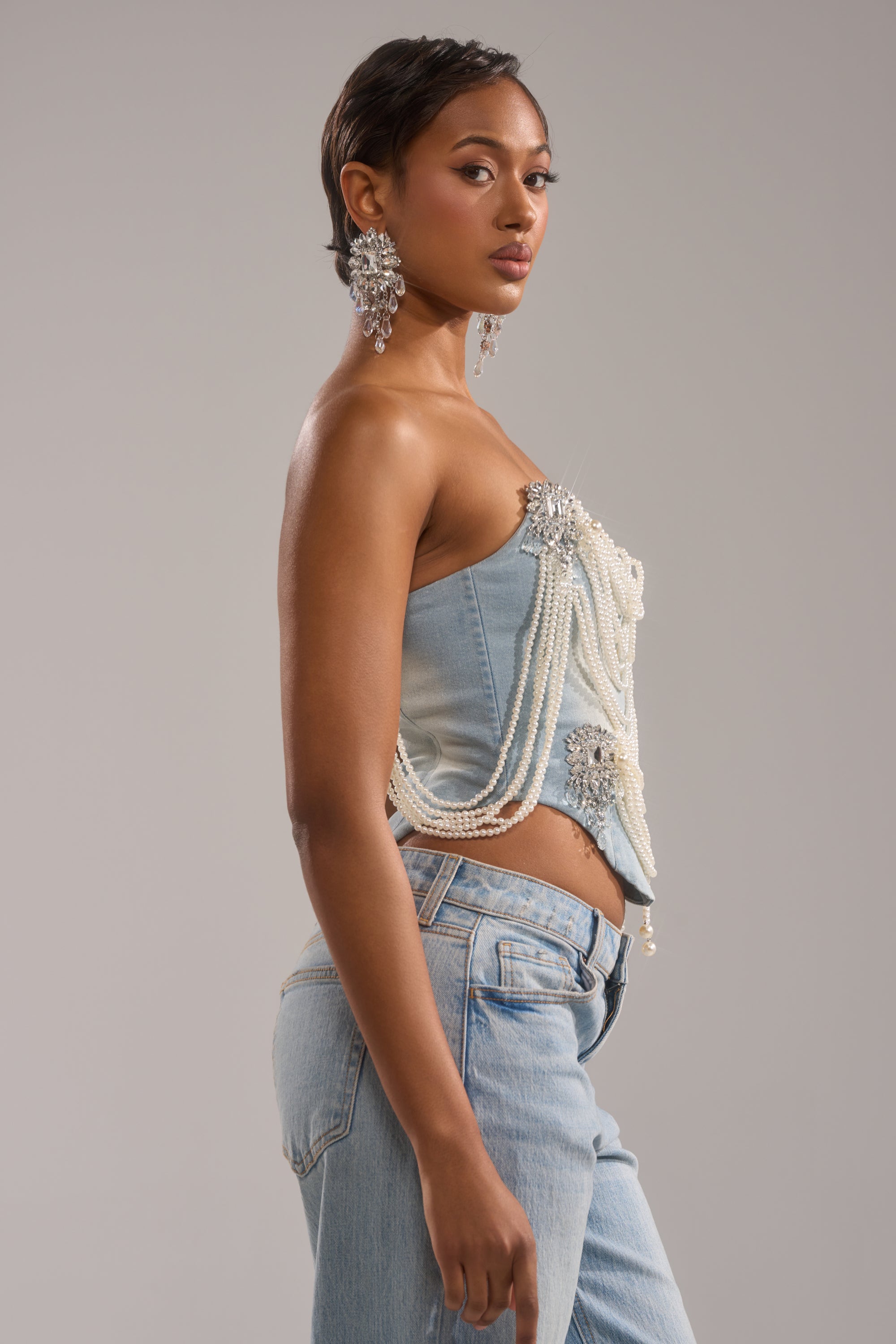 A woman in profile wears the PEARL QUEEN DENIM CORSET with pearl strands and brooches, paired with light blue jeans and large ornate earrings, standing against a neutral background.