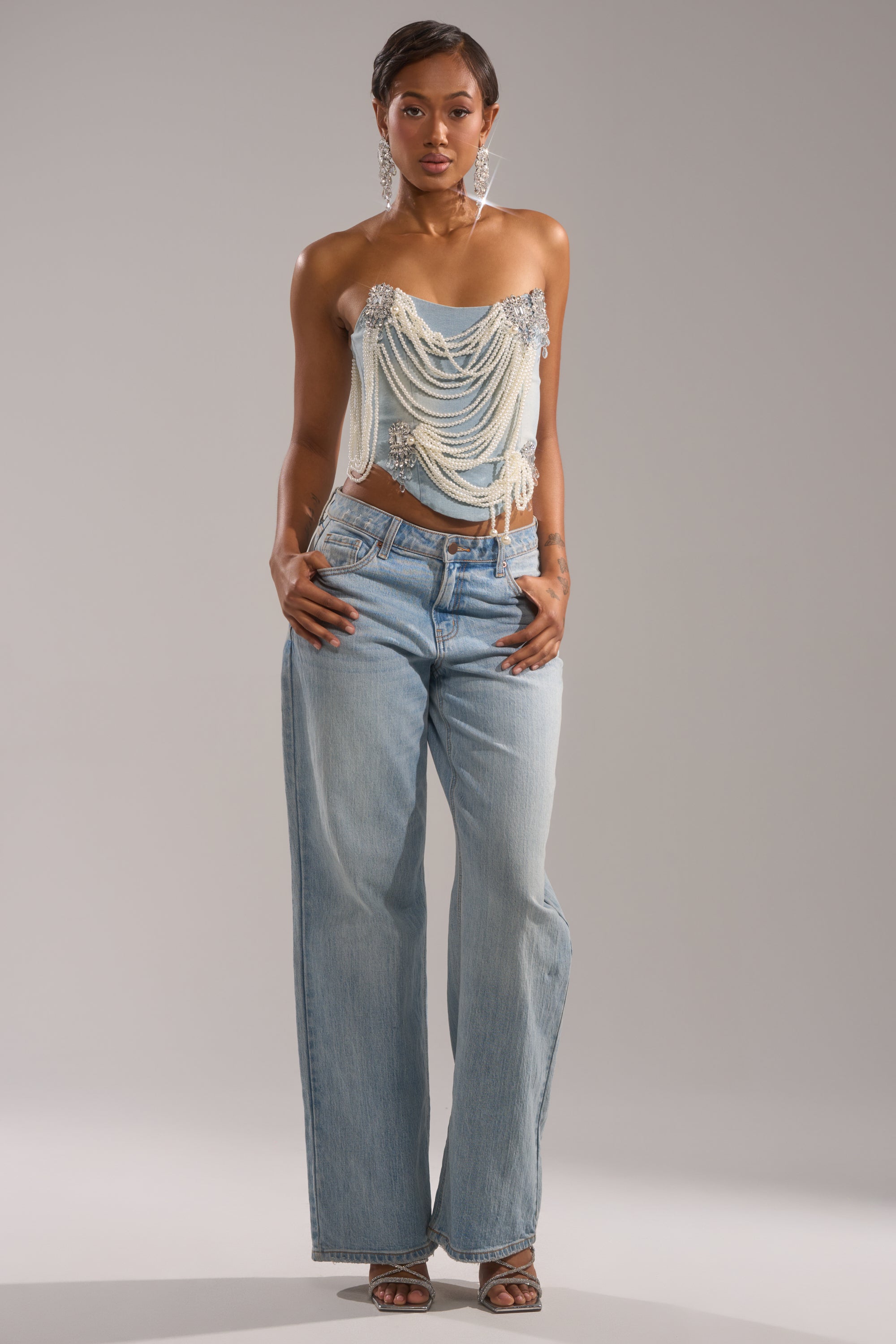 A woman faces forward wearing the PEARL QUEEN DENIM CORSET and light blue wide-leg jeans. She has short hair, statement earrings, and poses with her hands in her pockets against a plain background.