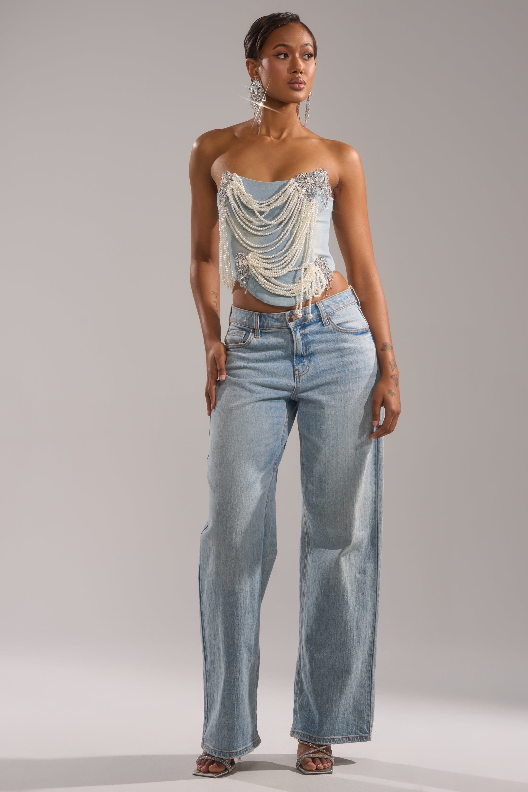 A woman wears the PEARL QUEEN DENIM CORSET with light blue wide-leg jeans, silver earrings, and strappy heels, posing against a neutral background.