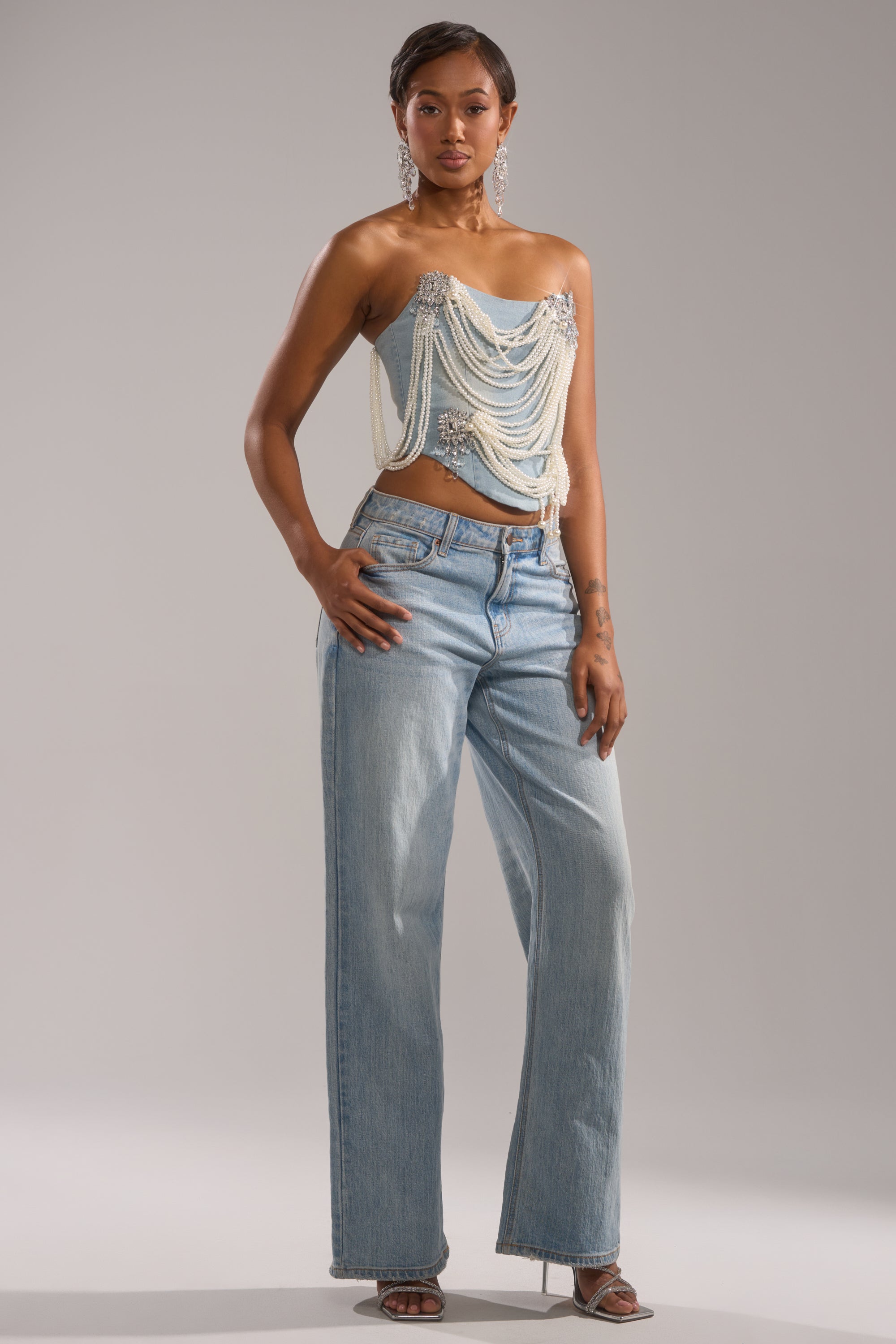 A woman poses in light blue wide-leg jeans, heeled sandals, and the PEARL QUEEN DENIM CORSET, featuring draped pearls and sparkling embellishments. She accessorizes with dangling earrings against a plain gray background.
