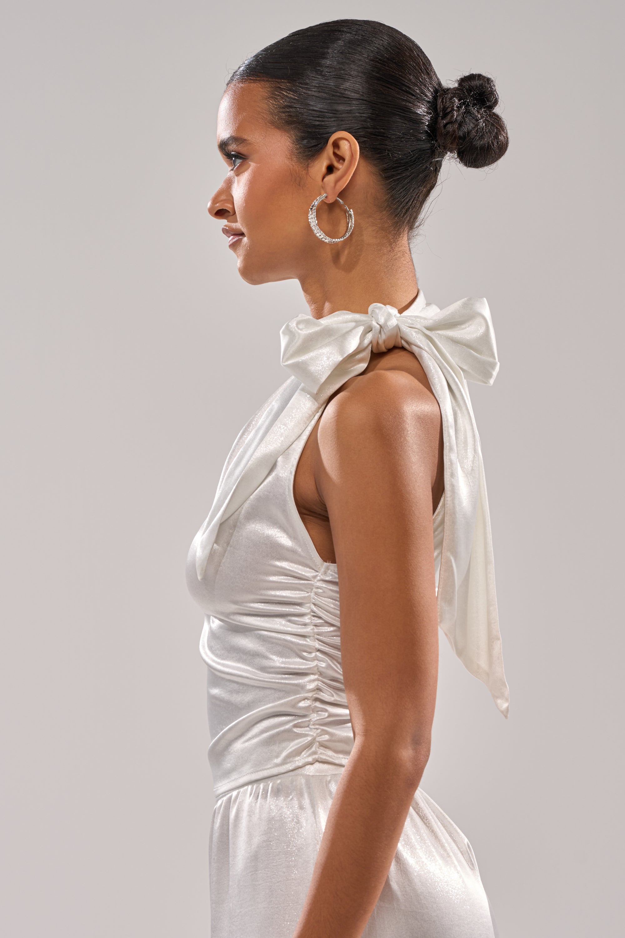 A woman in profile with a sleek bun and hoop earrings models the AKIRA Label LEAVE ME ALONE SLINKY METALLIC BLOUSE, which features a large shoulder bow, against a plain light background. 4