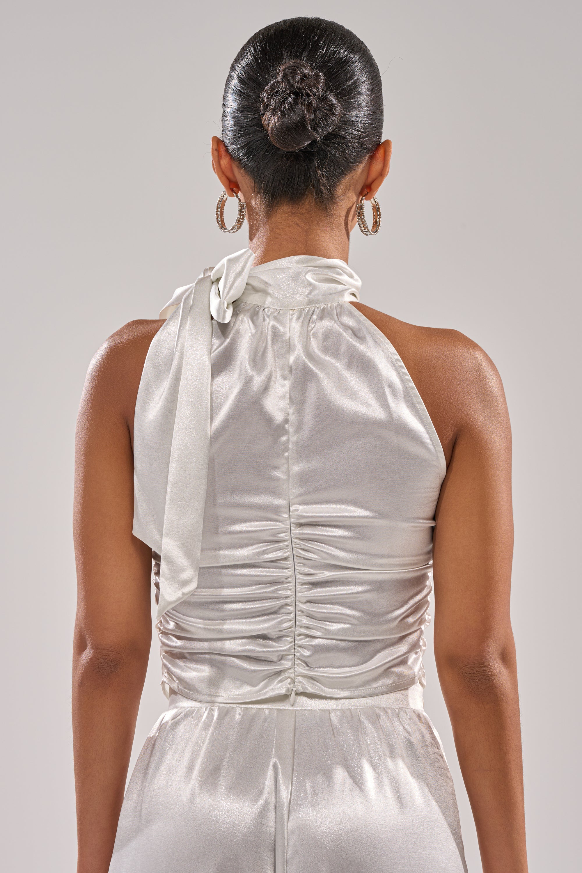 A woman with a sleek bun is shown from the back, wearing the LEAVE ME ALONE SLINKY METALLIC BLOUSE featuring a halter neckline, ruched detailing, and neck bow, paired with matching pants and large hoop earrings. 6
