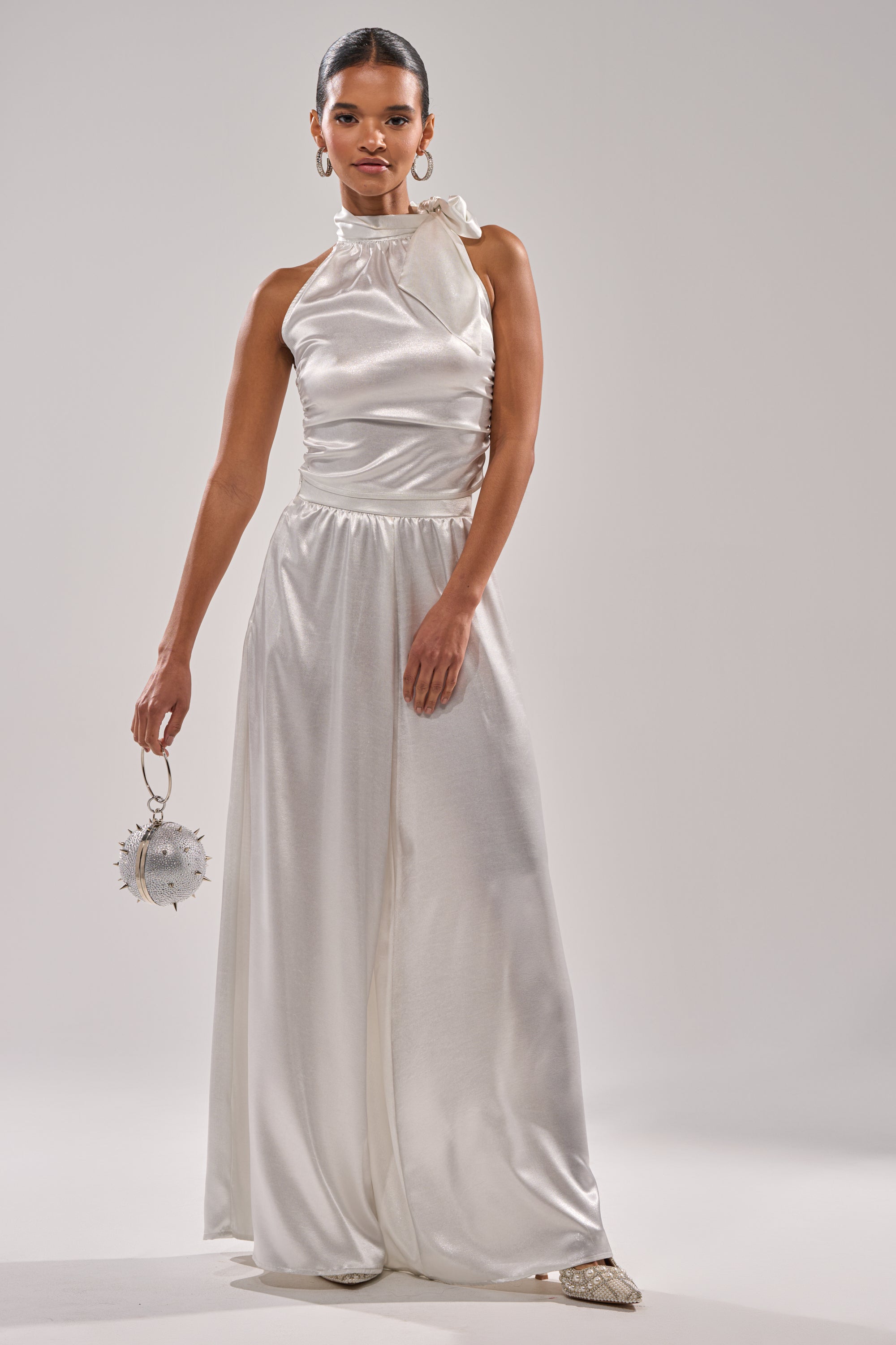 A woman stands confidently in the LEAVE ME ALONE SLINKY METALLIC BLOUSE, its metallic sheen catching the light. She holds a small, round embellished purse, pairs it with pointed shoes, and poses against a plain, light background. 7