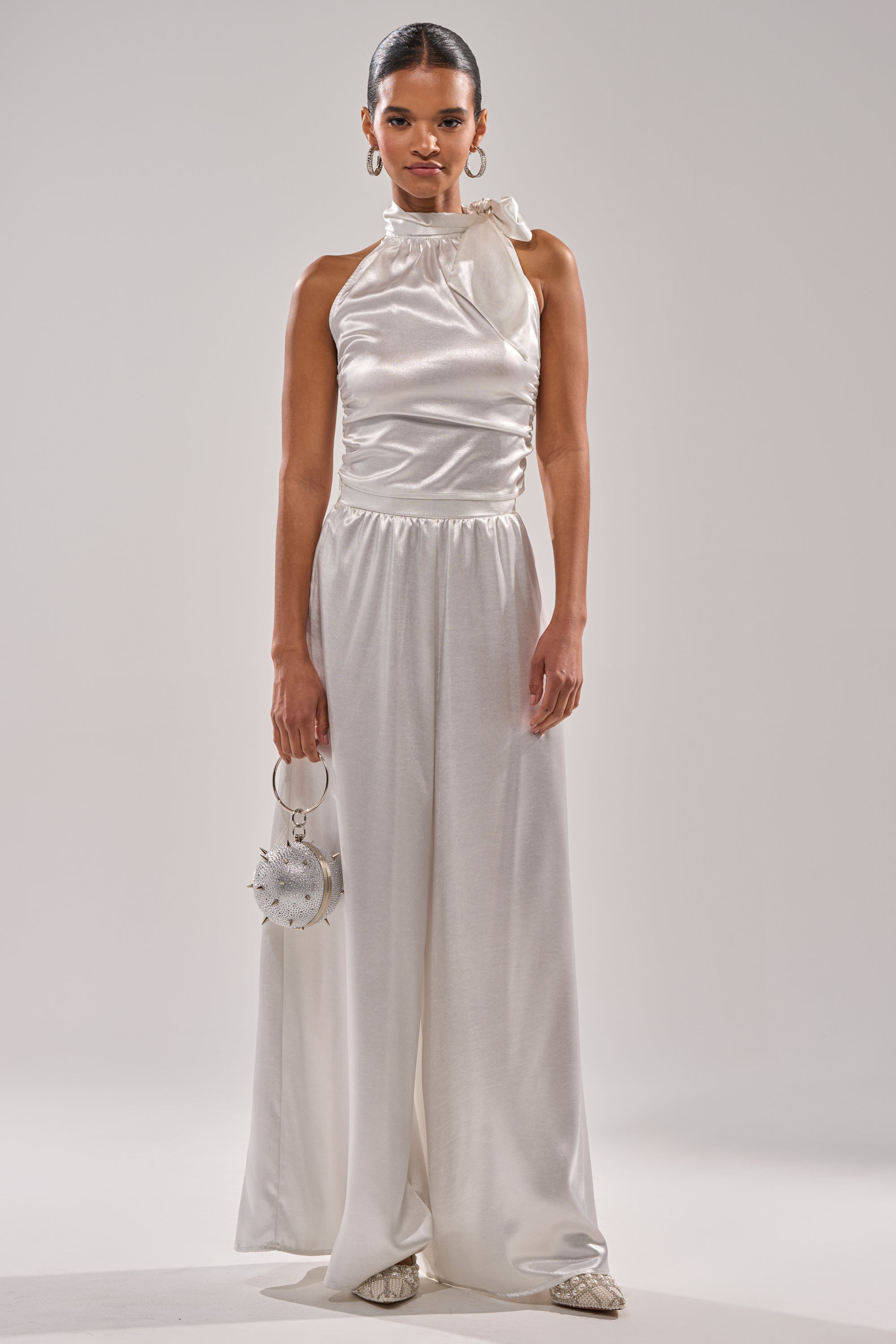 A woman in the LEAVE ME ALONE SLINKY METALLIC BLOUSE with hoop earrings and a round silver handbag stands against a plain light background—an ideal outfit for holiday events. 8
