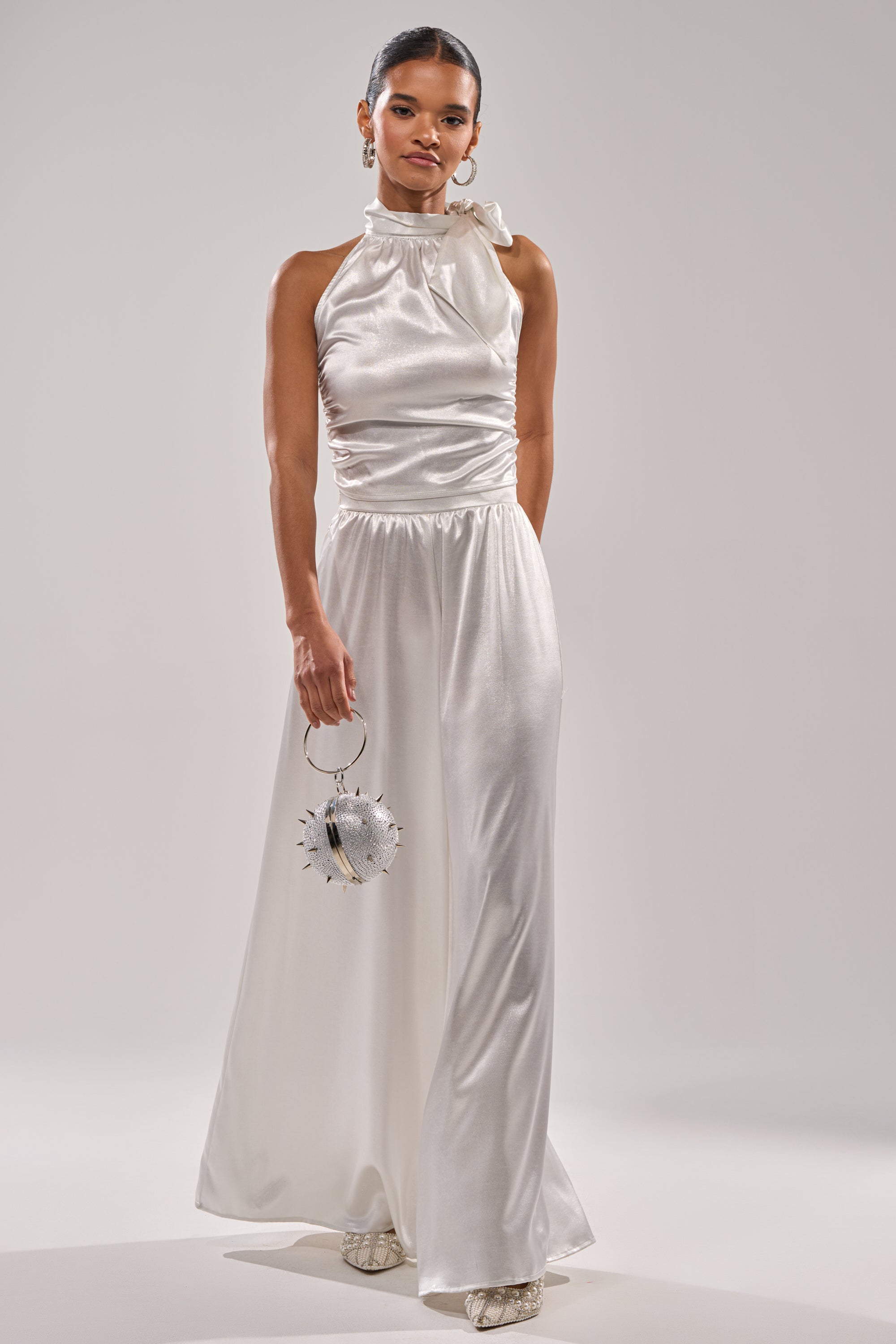 A woman in the LEAVE ME ALONE SLINKY METALLIC BLOUSE, featuring a sleeveless halter neckline, pairs it with a round silver spiked handbag—an ideal look for holiday events—standing against a plain light background. 9