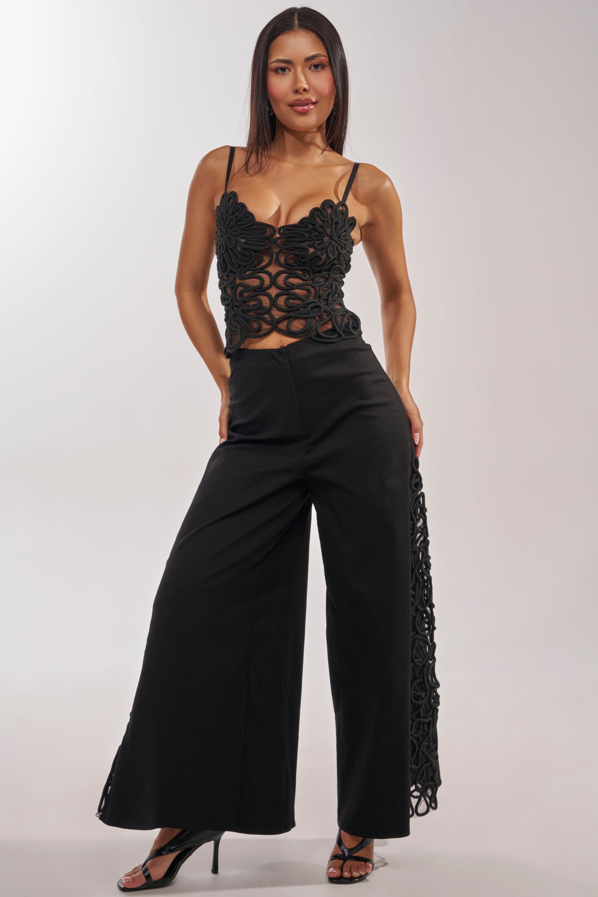 A woman stands with hands on hips, wearing the ITS GIVING ROPE DETAIL TROUSER paired with black high-heeled sandals and a sleeveless cutout black top, posing confidently against a plain background. 1