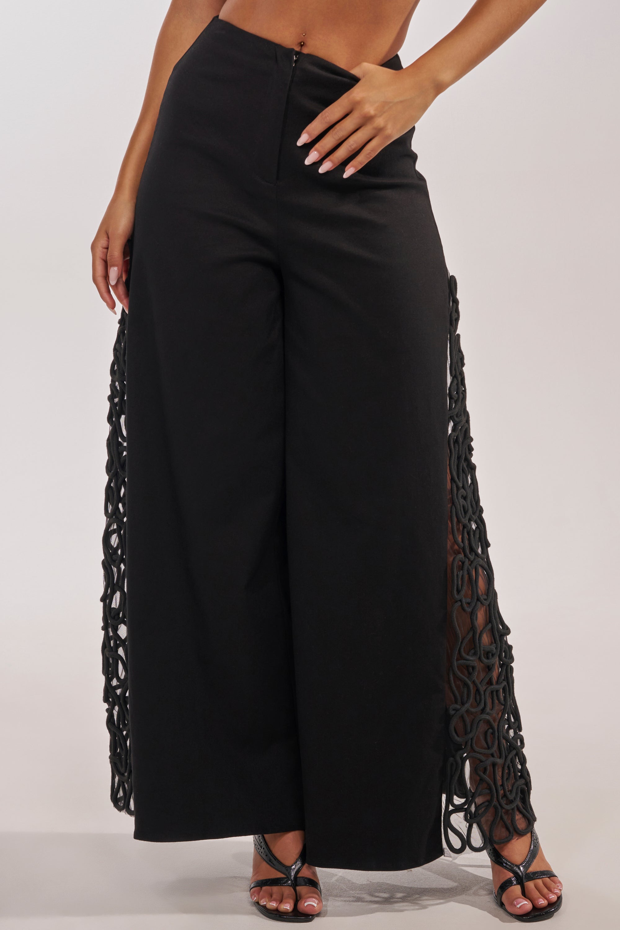A woman wears the ITS GIVING ROPE DETAIL TROUSER, featuring high-waisted wide-leg design with rope accents, ideal for formal occasions. She pairs them with open-toe heels and her hand rests on her hip, showing off light pink nail polish. 2