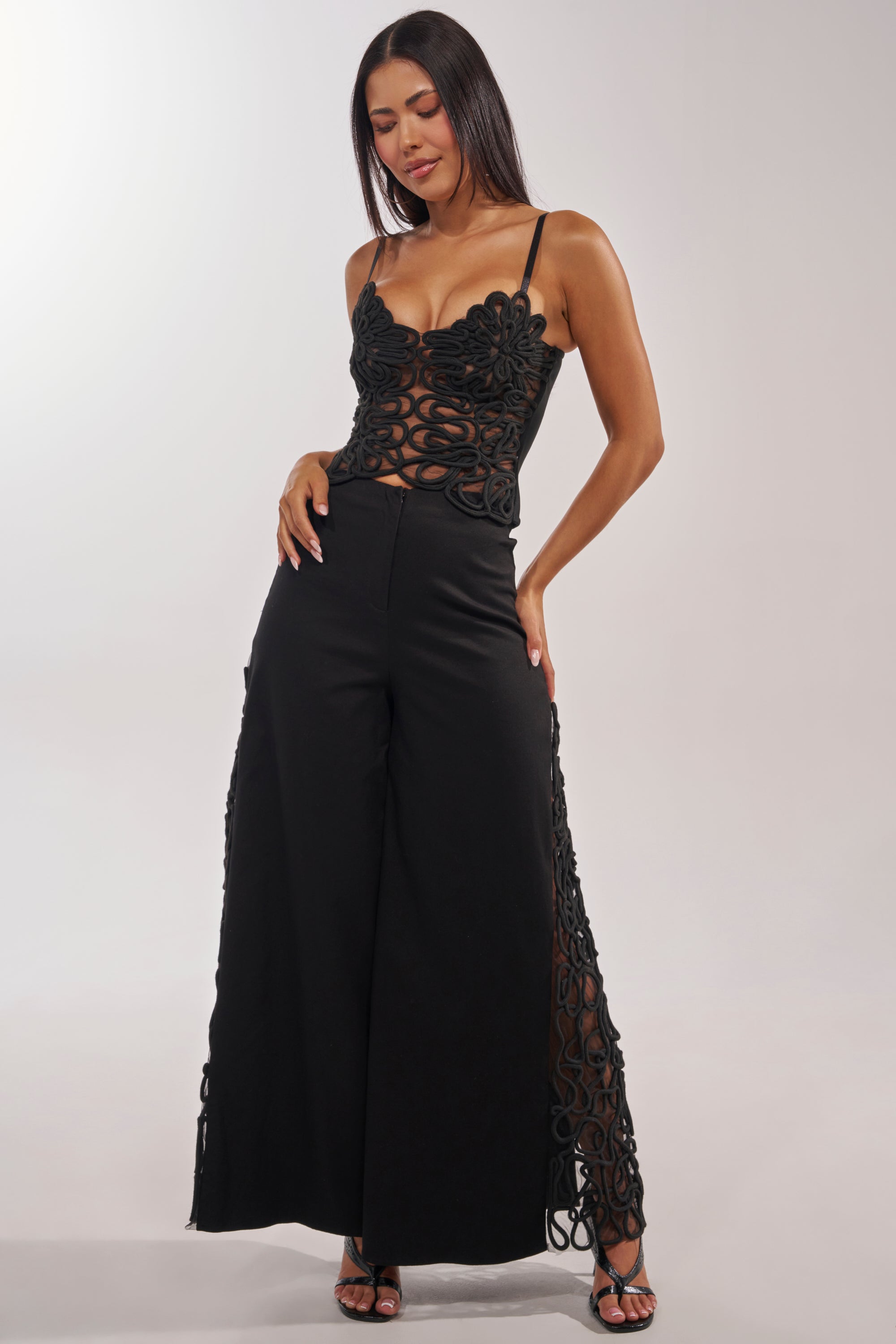 A woman wears the ITS GIVING ROPE DETAIL TROUSER, a pair of high-waisted, wide-leg black pants with intricate rope and sheer cut-out details, styled with strappy black heels against a plain background. 0