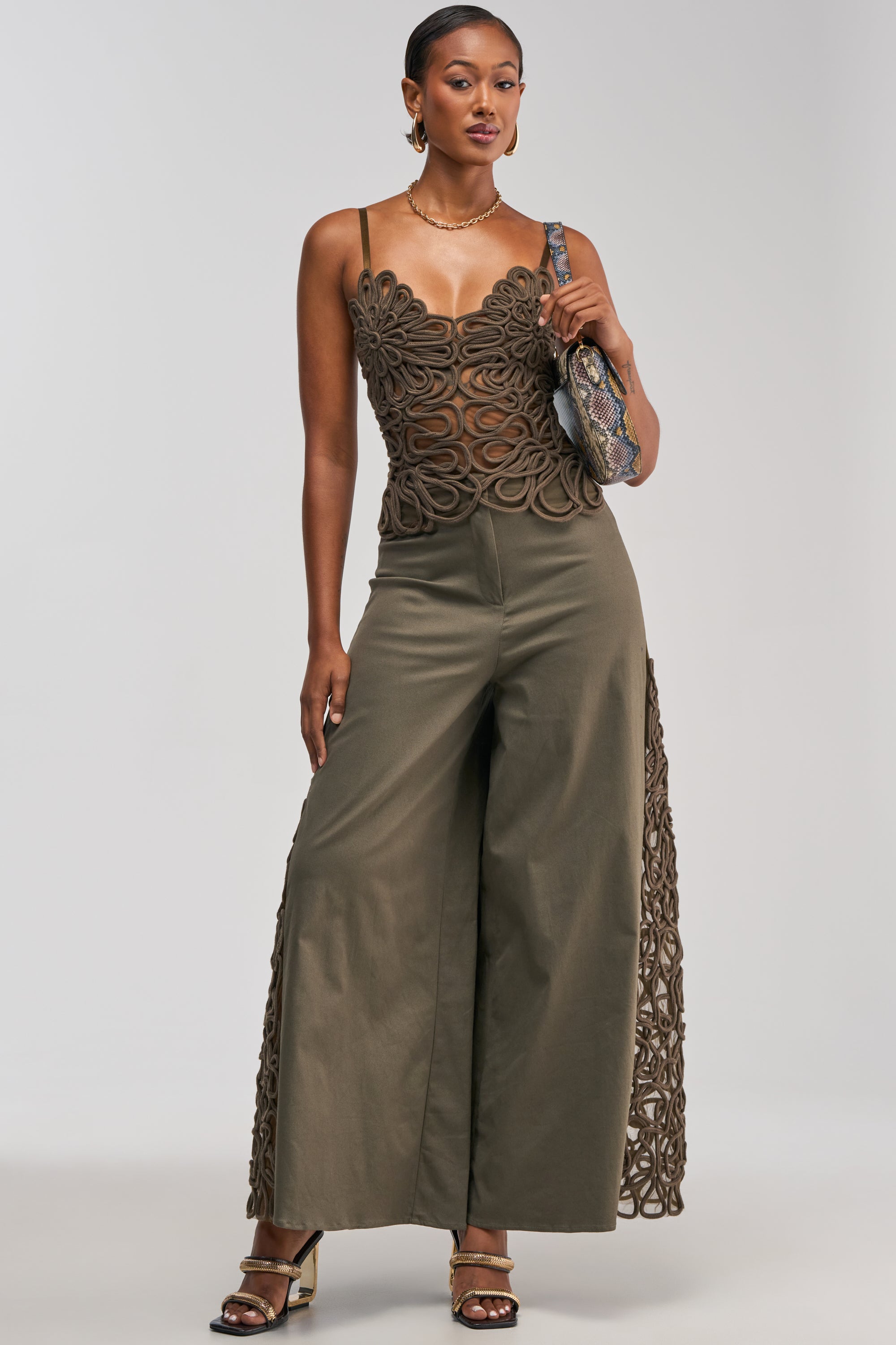 A woman models the ITS GIVING Rope Detail Trouser in olive, styled with a brown embroidered sleeveless top, openwork and rope accents, gold earrings, a necklace, strappy heels, and a textured shoulder bag. She stands against a plain background for an effortlessly chic business-casual look.