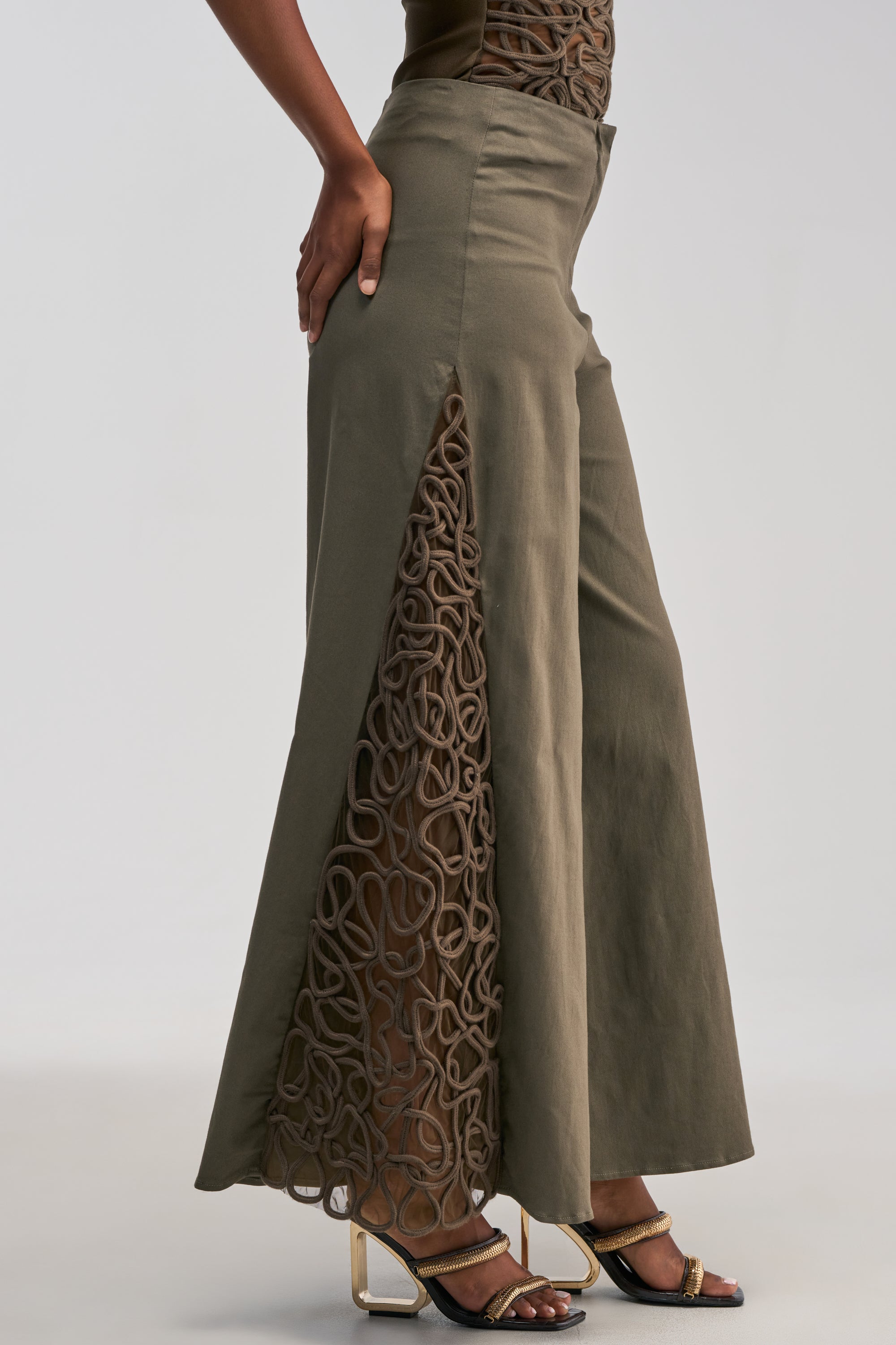 Wearing the ITS GIVING ROPE DETAIL TROUSER IN OLIVE—wide-leg trousers with side mesh cutouts showcasing intricate swirl patterns and subtle rope detailing—paired with heeled sandals.