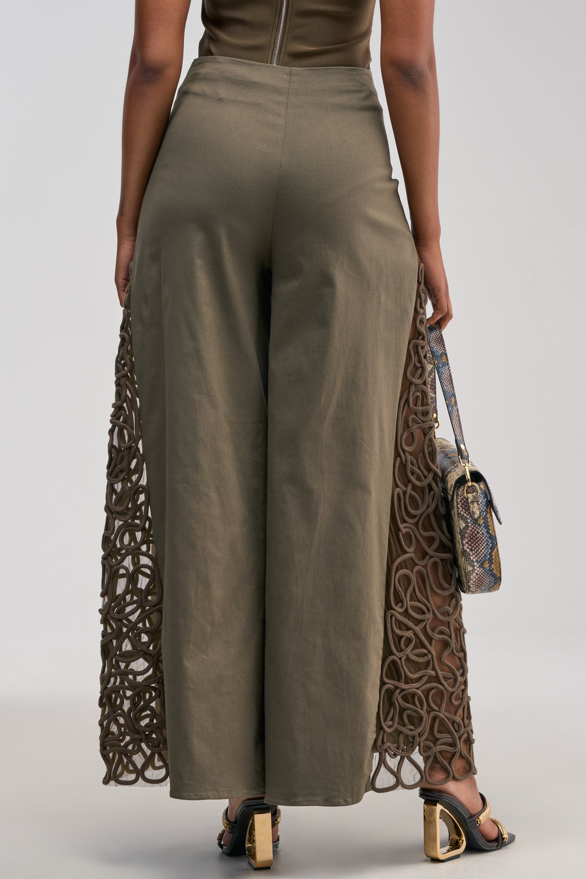 A person stands facing away, wearing the ITS GIVING ROPE DETAIL TROUSER IN OLIVE—wide-leg trousers featuring intricate lace-like cutouts and subtle rope detailing on the sides—paired with heeled sandals and a snakeskin handbag.