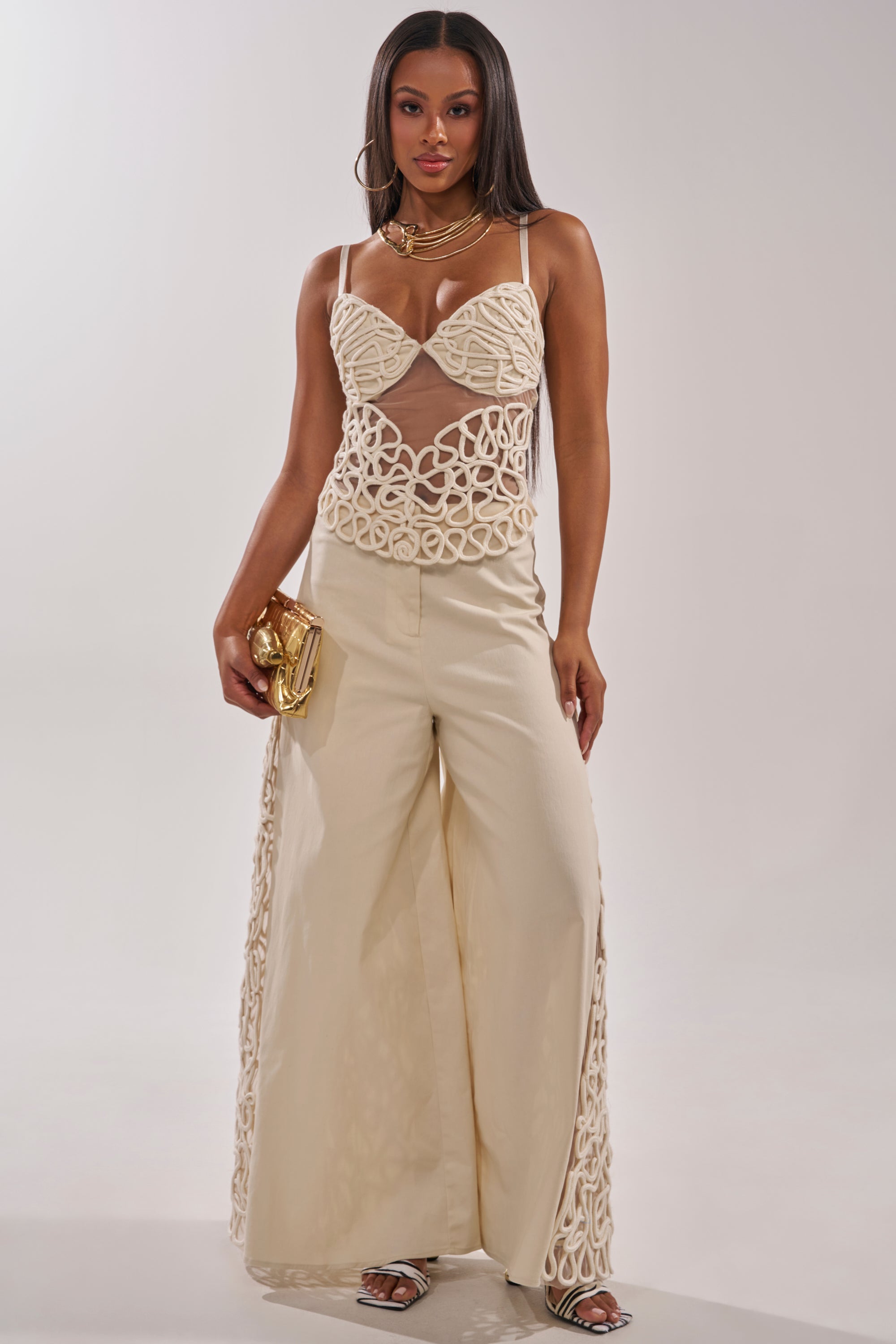 A woman stands in a white studio wearing the NEVER LOSE ME ROPE CORSET, styled with wide-leg cream pants. She holds a gold clutch and accessorizes with layered necklaces, hoop earrings, and heeled sandals. 5