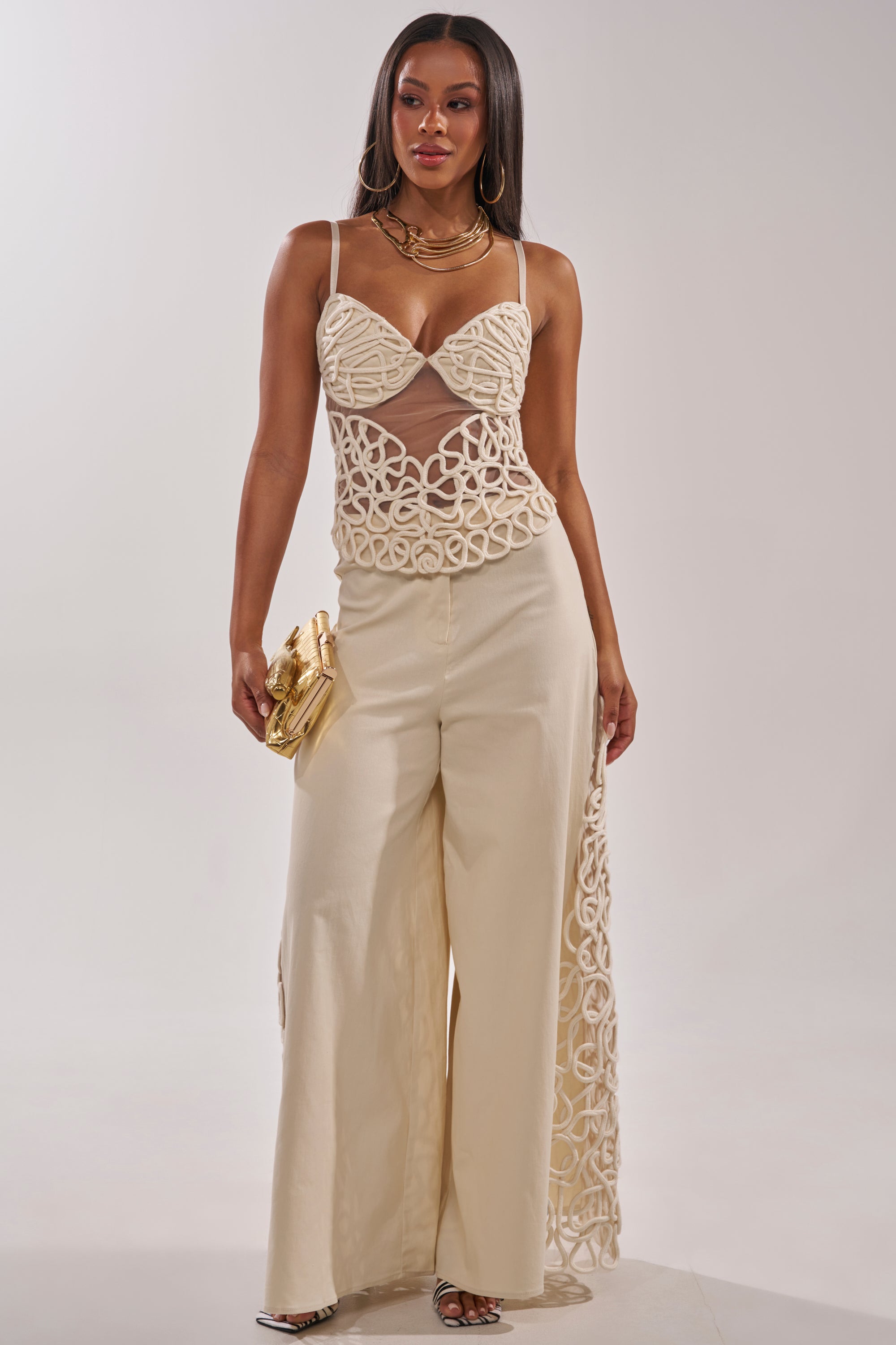 A woman in a stylish cream outfit poses against a plain background, wearing wide-leg pants and the NEVER LOSE ME ROPE CORSET. She accessorizes with layered gold necklaces, a gold clutch, and strappy heels. 6