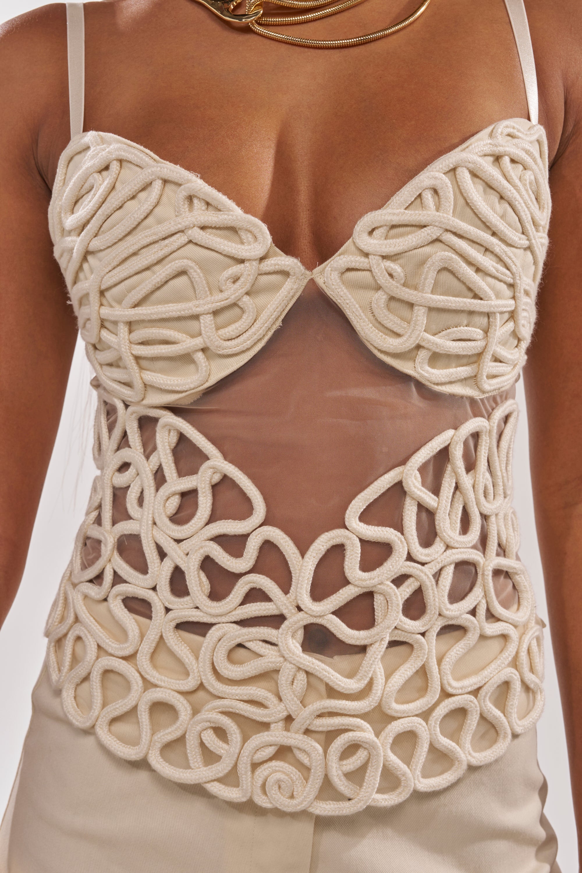 A close-up of a woman's torso wearing the NEVER LOSE ME ROPE CORSET, a sheer mesh top with intricate cream rope detailing and spaghetti straps, paired with cream pants and a gold necklace. 8