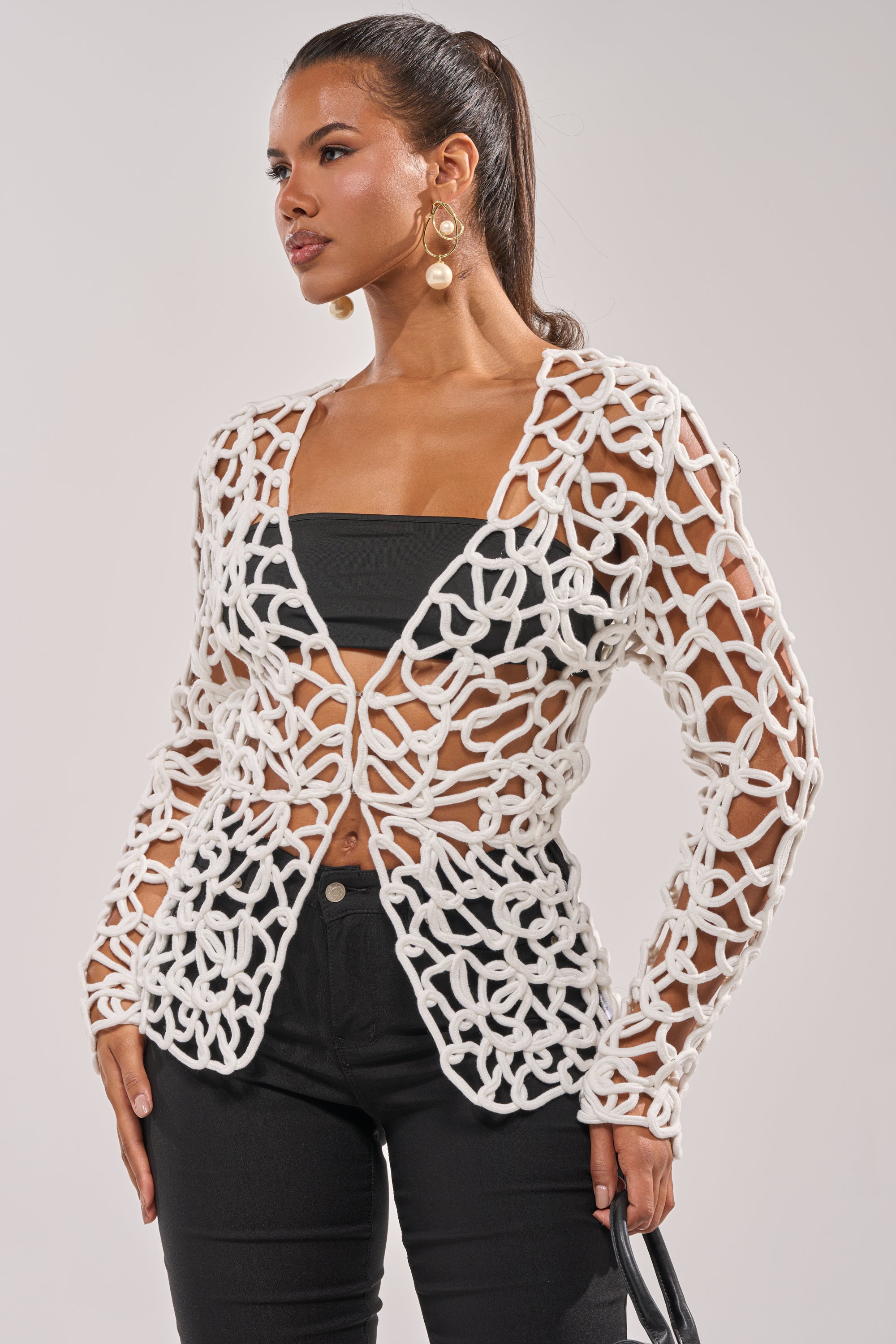 A woman poses against a plain light background, wearing the LIKE YOU'VE NEVER SEEN CROCHET BLOUSE over a black bandeau and pants. Large gold earrings complete her look, making the open-front blouse stand out stylishly. 0
