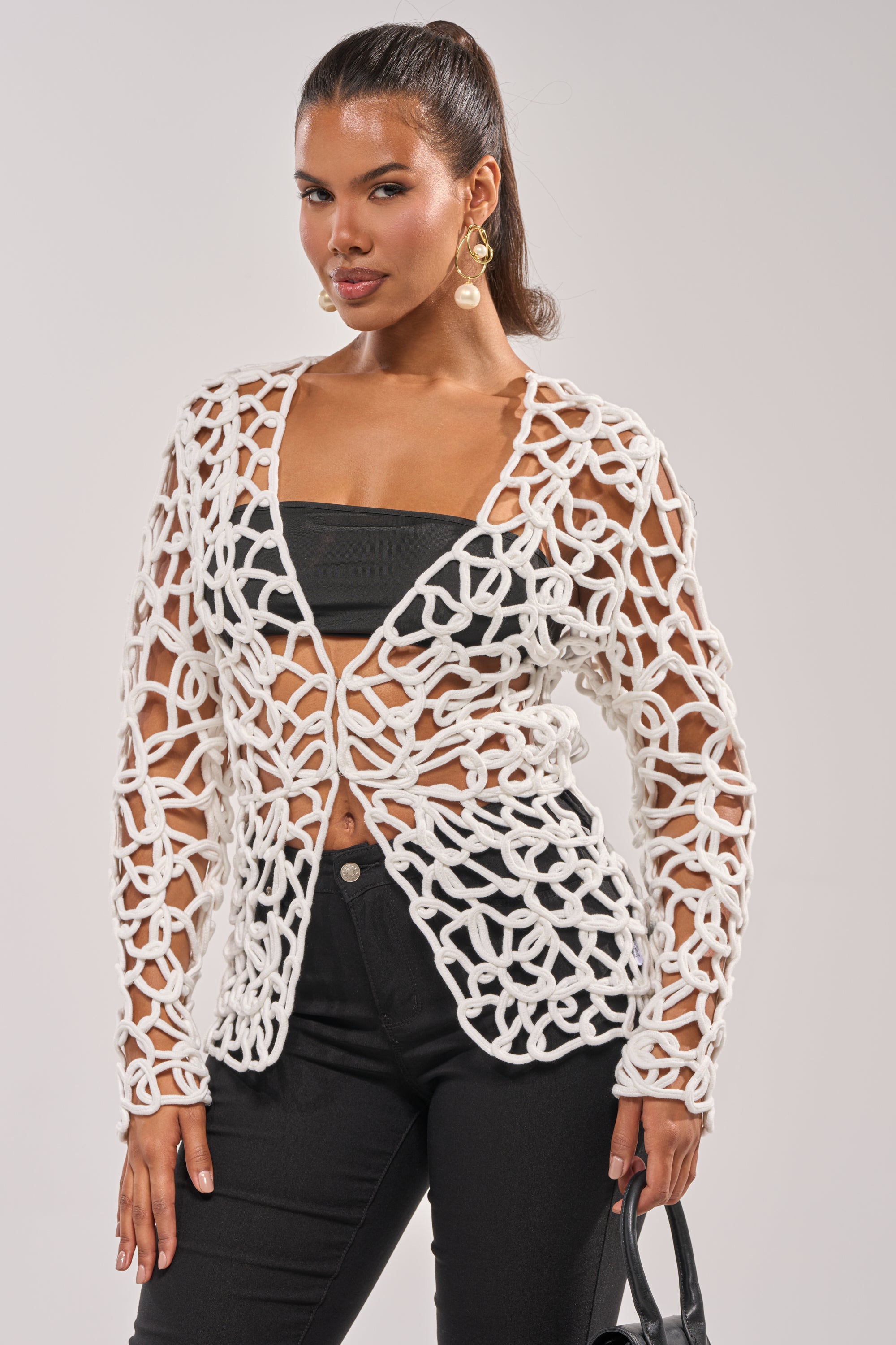 A woman poses against a light background wearing the LIKE YOU'VE NEVER SEEN CROCHET BLOUSE open over a black tube top and pants, accessorized with dangling earrings and a black handbag. 1