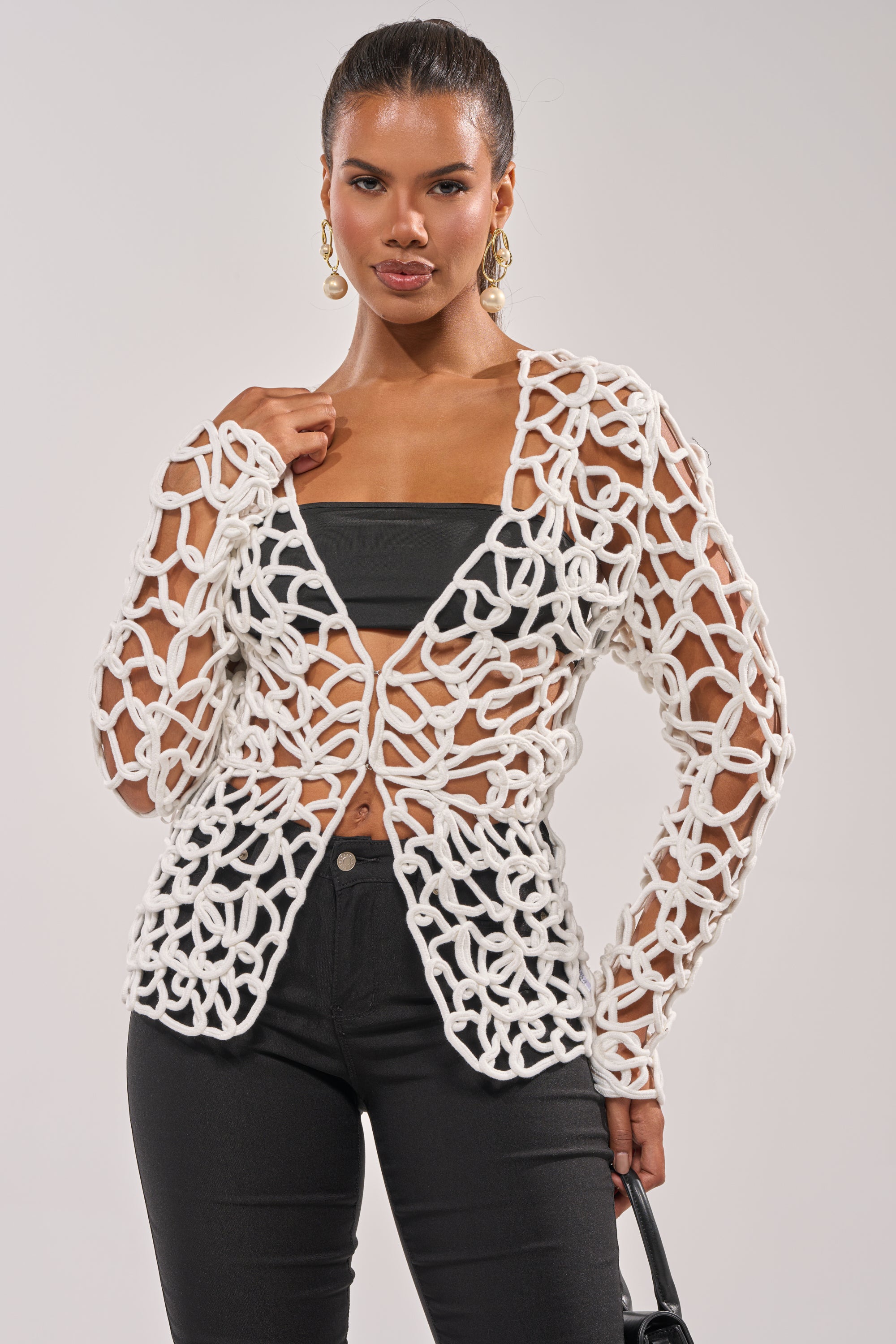 A woman models the LIKE YOU'VE NEVER SEEN CROCHET BLOUSE, a chunky, open-knit white top with a geometric pattern layered over a black tube top and high-waisted pants. She finishes the look with gold earrings and a black handbag. 2