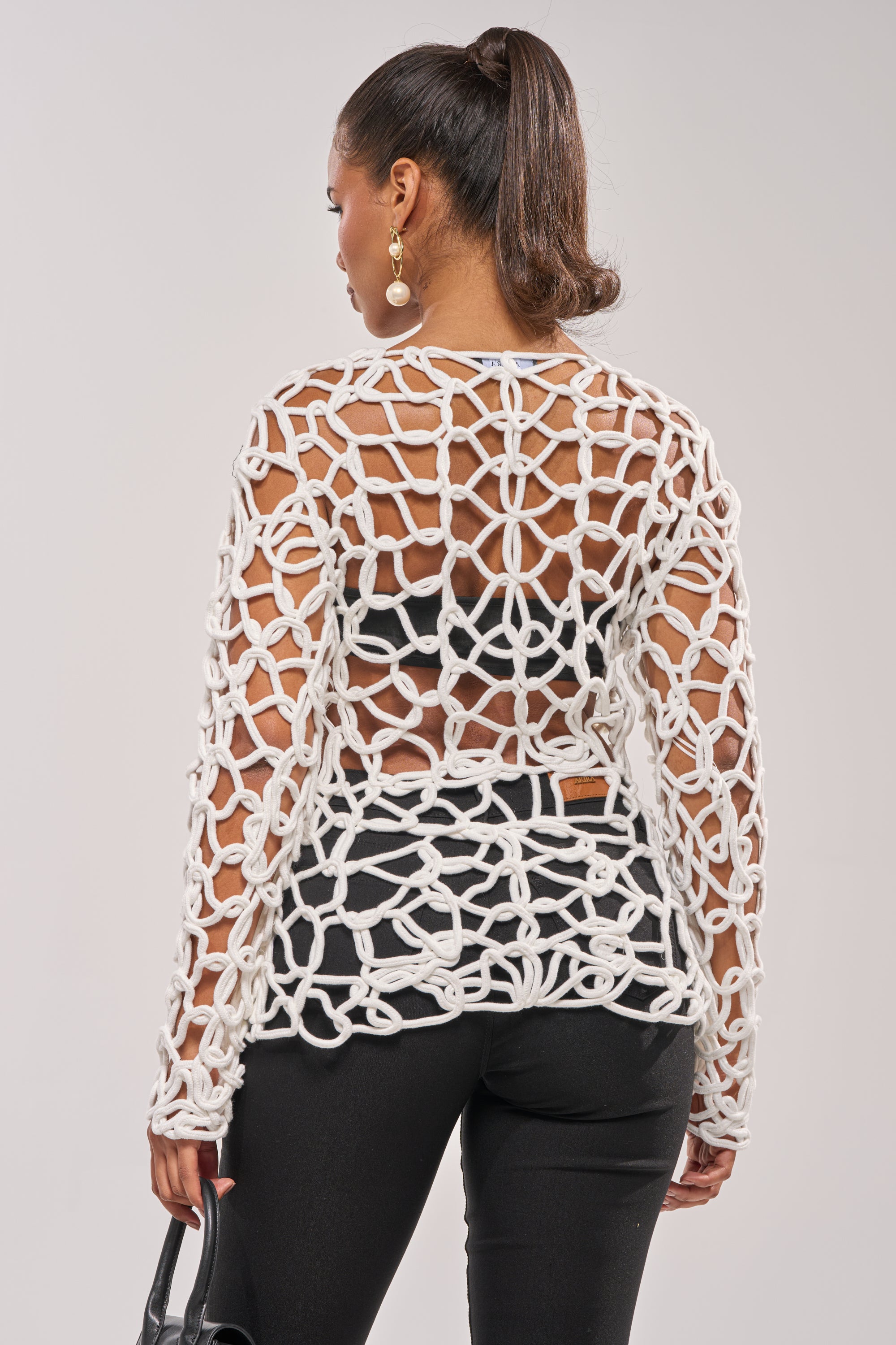 A woman faces away, wearing the LIKE YOU'VE NEVER SEEN CROCHET BLOUSE in white over a black top and pants. She carries a black handbag, her hair in a high ponytail, accessorized with large round earrings. 4