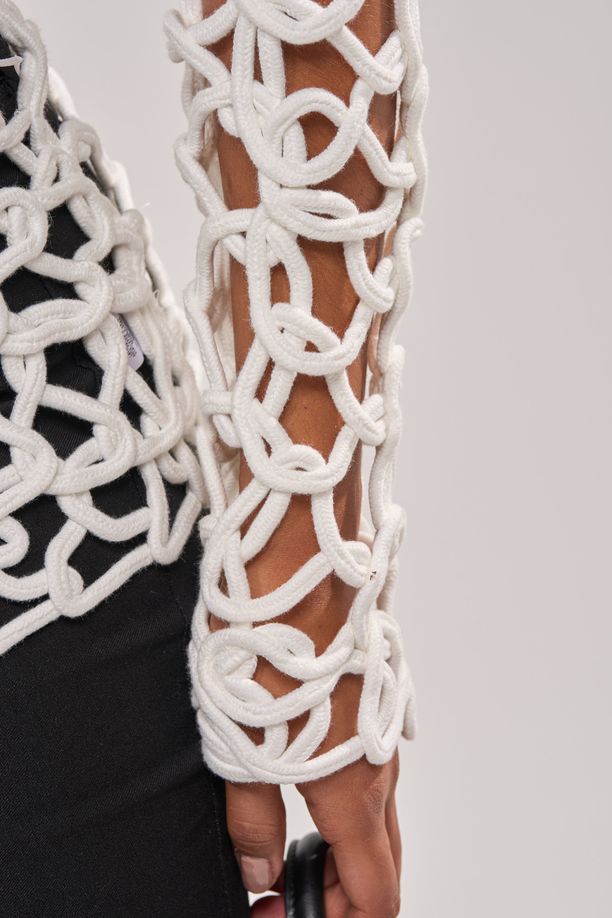 A close-up of someone wearing the LIKE YOU'VE NEVER SEEN CROCHET BLOUSE in white over a black top, showcasing the blouse’s detailed loops and knots on their arm. 6