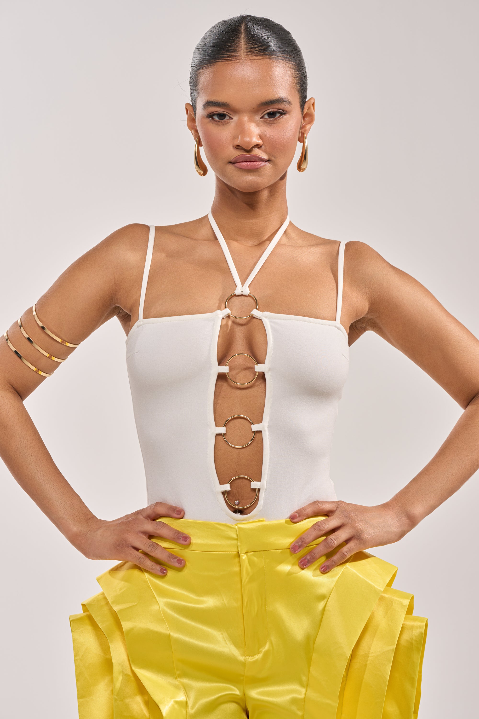 A woman with slicked-back hair wears the FLY ME OUT BODYSUIT featuring a white halter neckline and gold ring hardware, paired with bold gold jewelry and yellow pleated pants, standing confidently against a plain background. 0