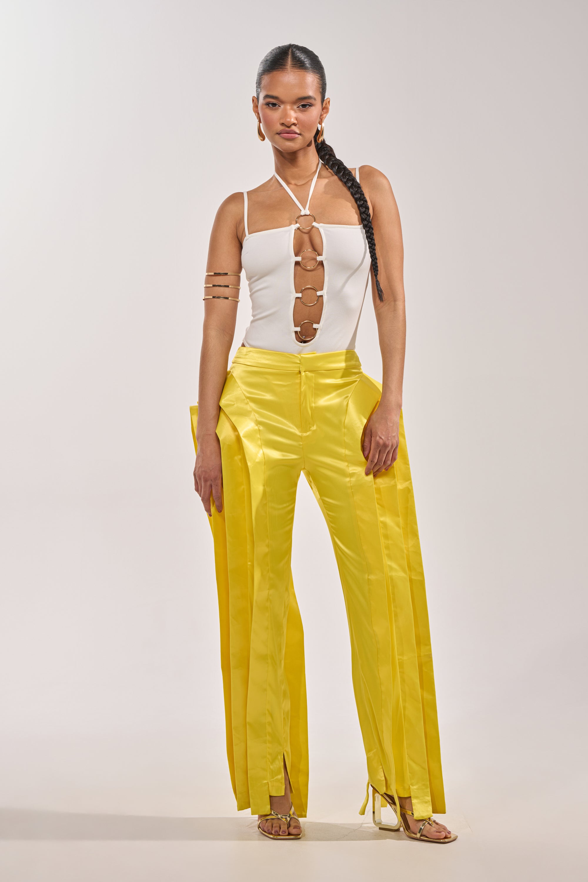 A woman poses confidently in the FLY ME OUT BODYSUIT, a white halter-neck piece with cut-out rings down the front, paired with bright yellow wide-leg pants, strappy heels, a long braid, and minimal accessories against a plain background. 4