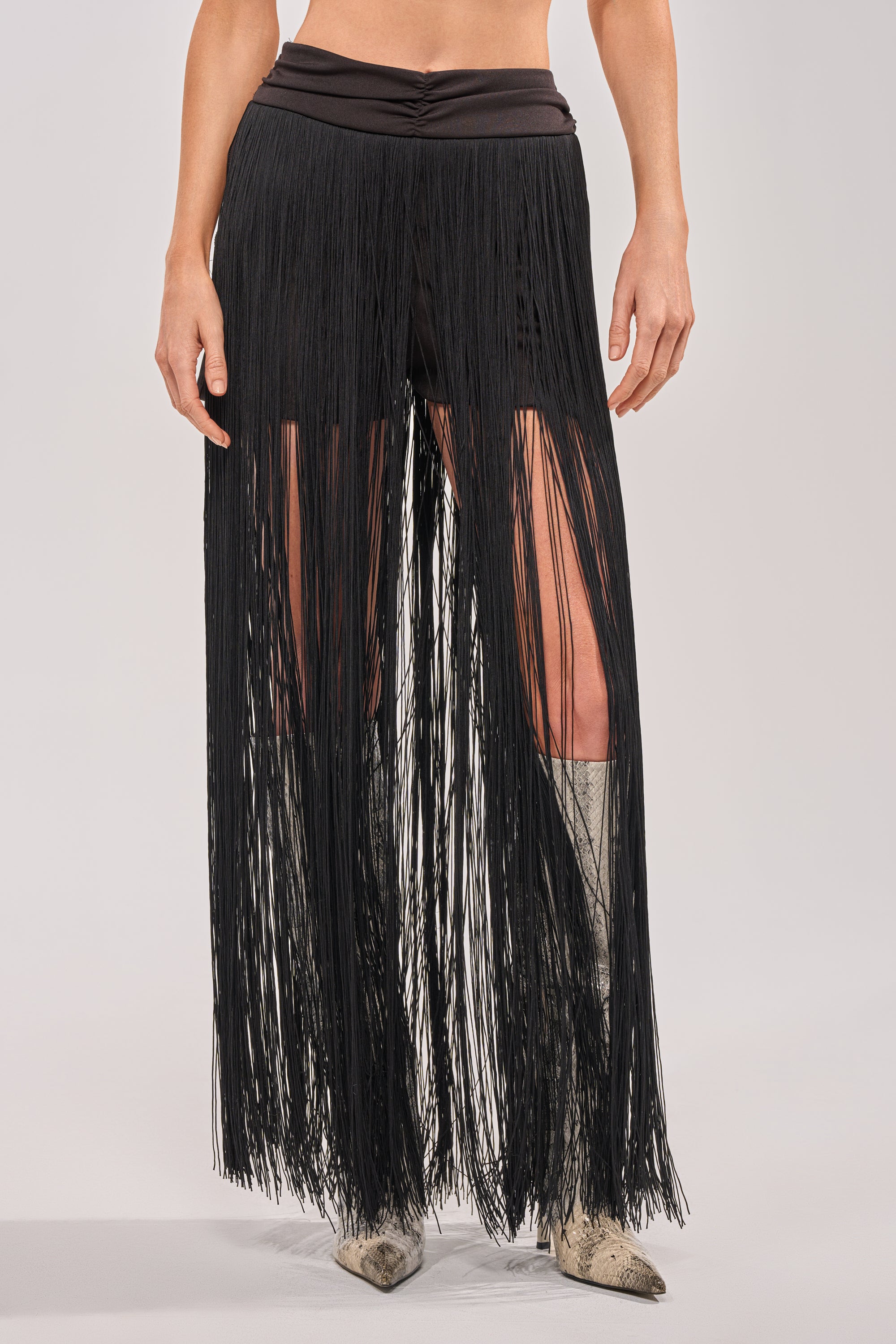 A person wearing the ON THE EDGE FRINGE SKIRT IN BLACK, featuring a ruched waistband and long black fringe, paired with silver pointed-toe boots. The image is cropped to show only the lower body. 0
