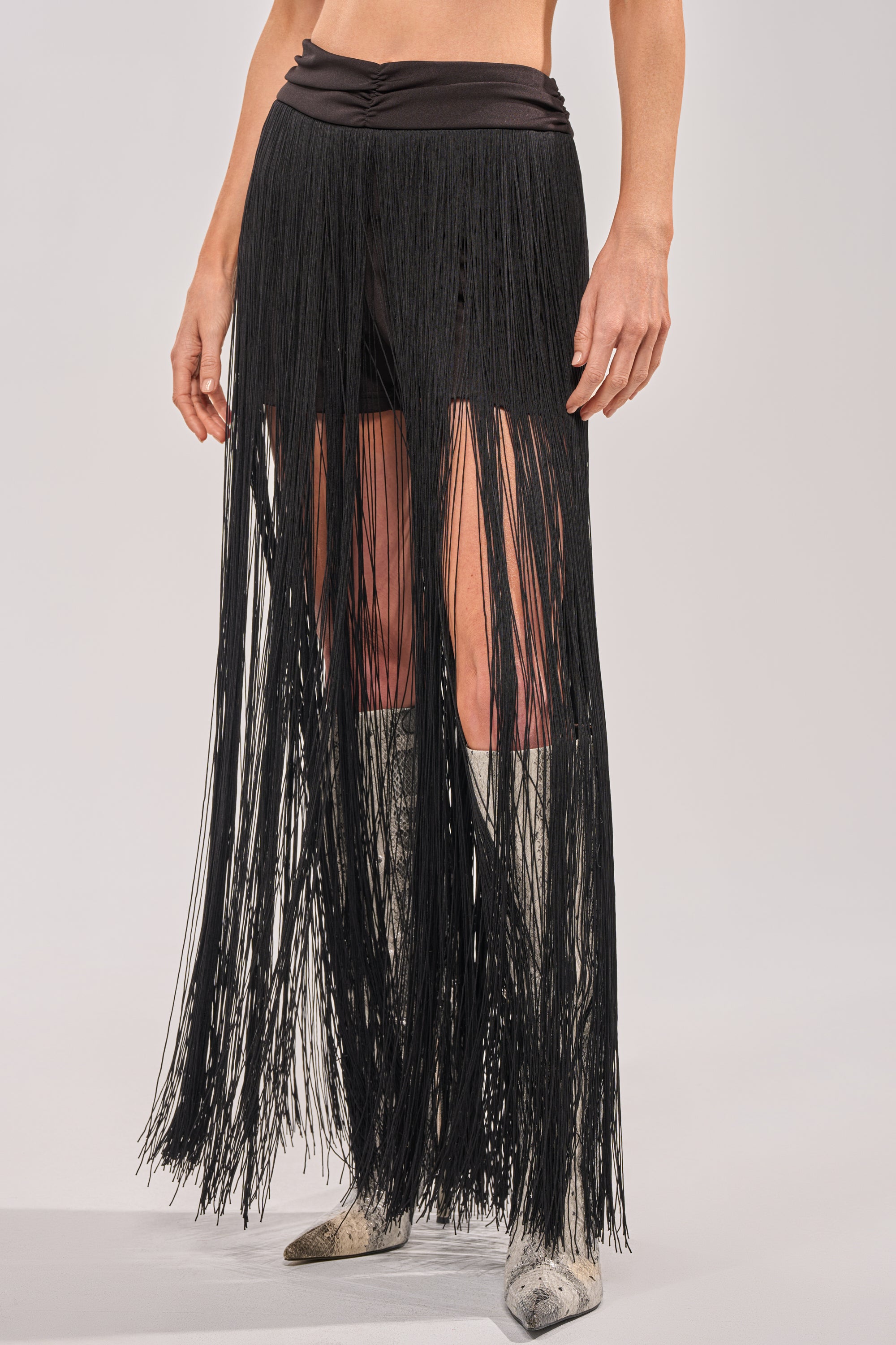 Wearing the ON THE EDGE FRINGE SKIRT IN BLACK featuring a ruched waistband over black shorts and silver pointed-toe boots, this ankle-length fringe skirt adds dramatic style for a party-ready look. 1