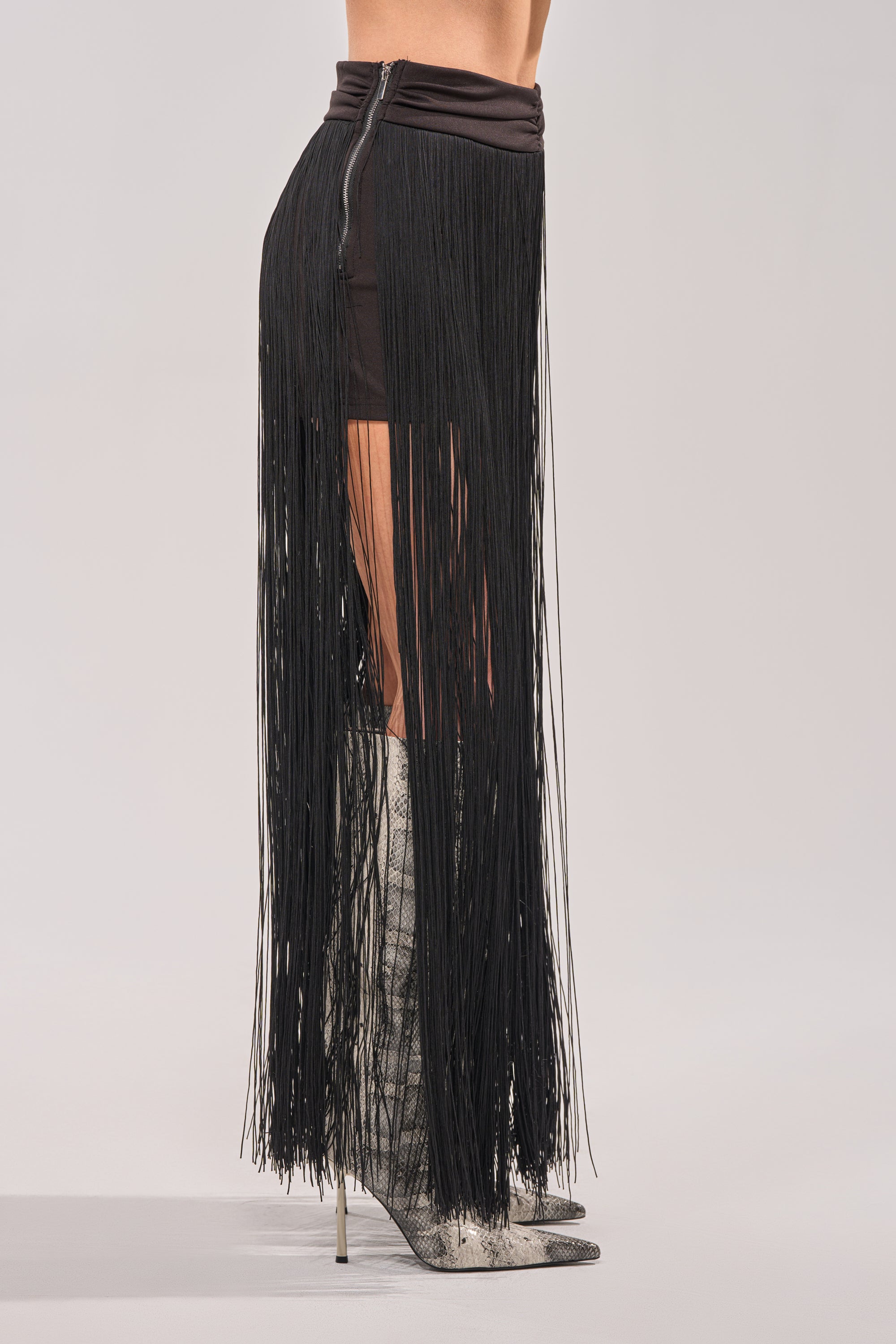 A person wears the ON THE EDGE FRINGE SKIRT IN BLACK, featuring long black fringe extending to the ankles, paired with knee-high pointed-toe snakeskin boots. The side-view image is set against a plain background. 2
