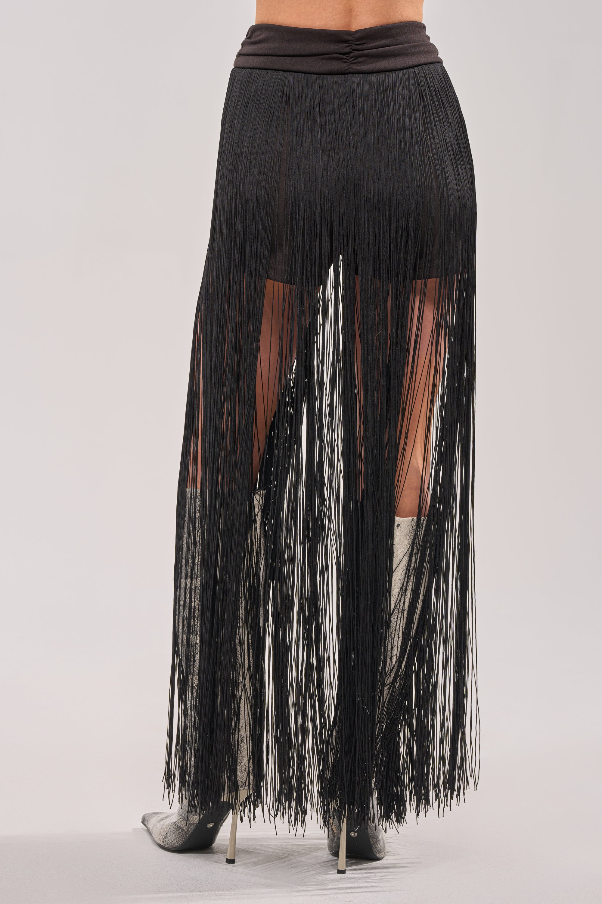 A person is shown from the back against a plain light background, wearing the ON THE EDGE FRINGE SKIRT IN BLACK with a ruched waistband and silver high-heeled boots. 3