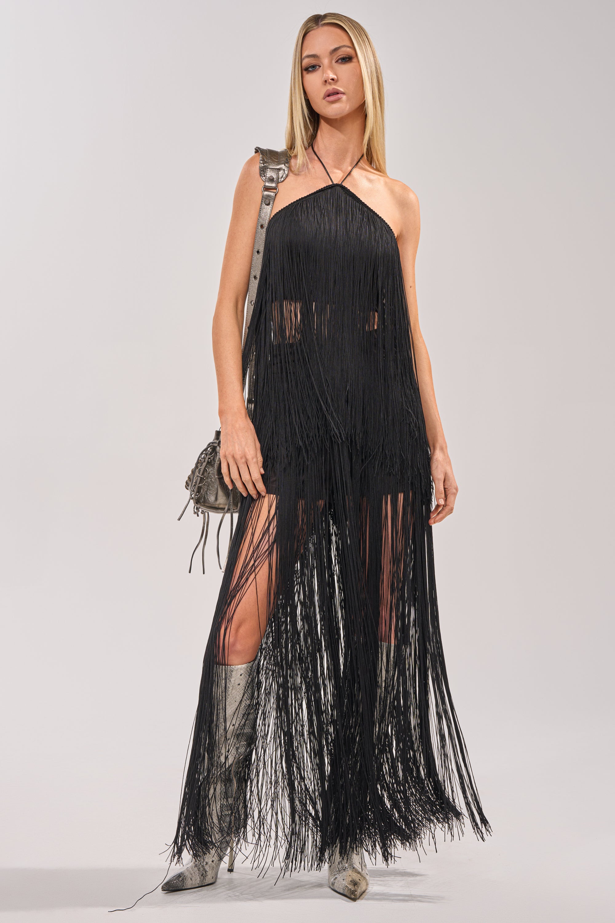 A woman wears the ON THE EDGE FRINGE HALTER TOP IN BLACK styled with knee-high silver boots and a small gray shoulder bag, posing against a plain light background. 0