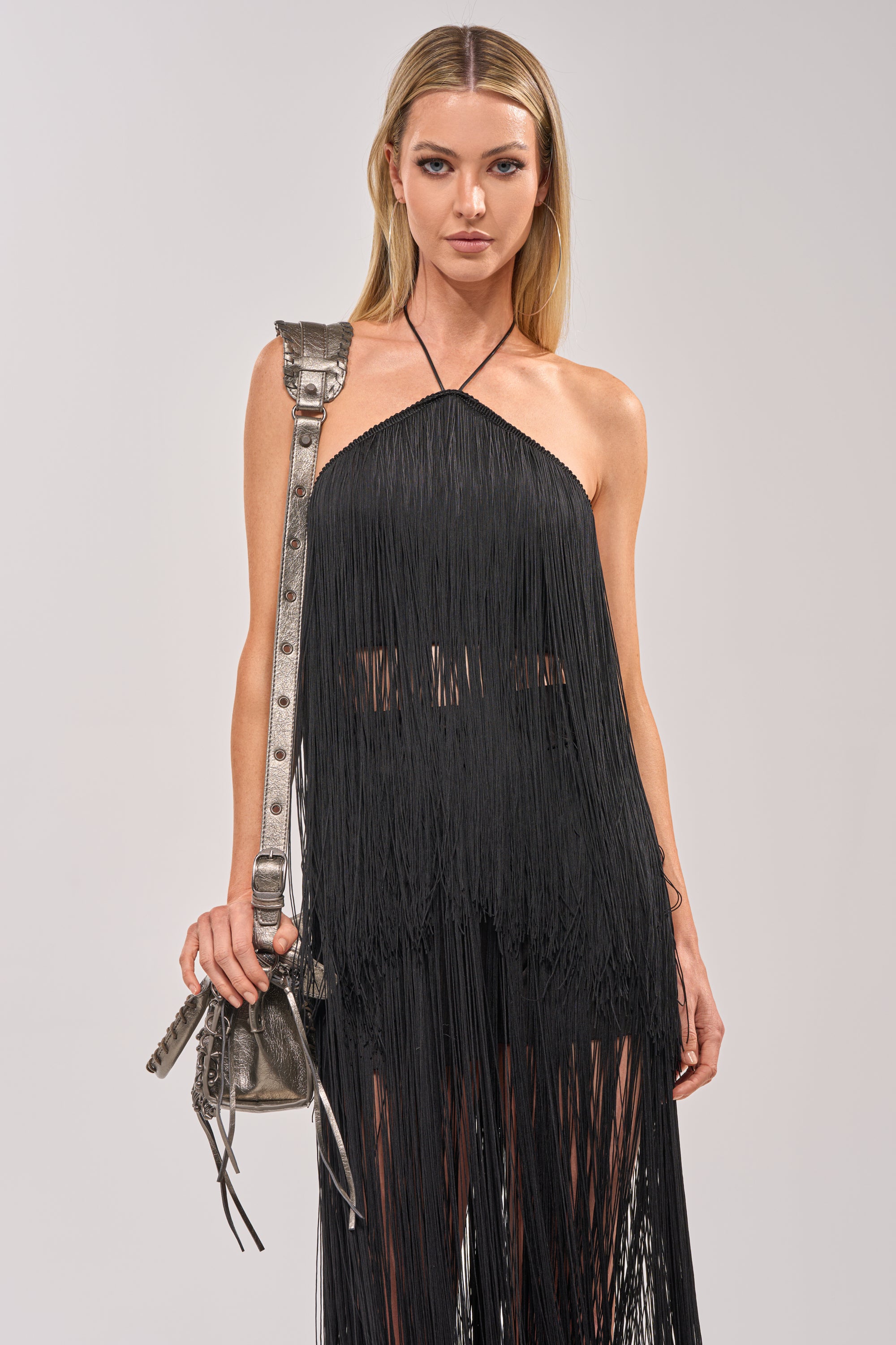 Perfect for a girls’ night out, a woman with long blonde hair wears the ON THE EDGE FRINGE HALTER TOP IN BLACK and carries a metallic shoulder bag. She stands against a plain light gray background, looking confidently at the camera. 2
