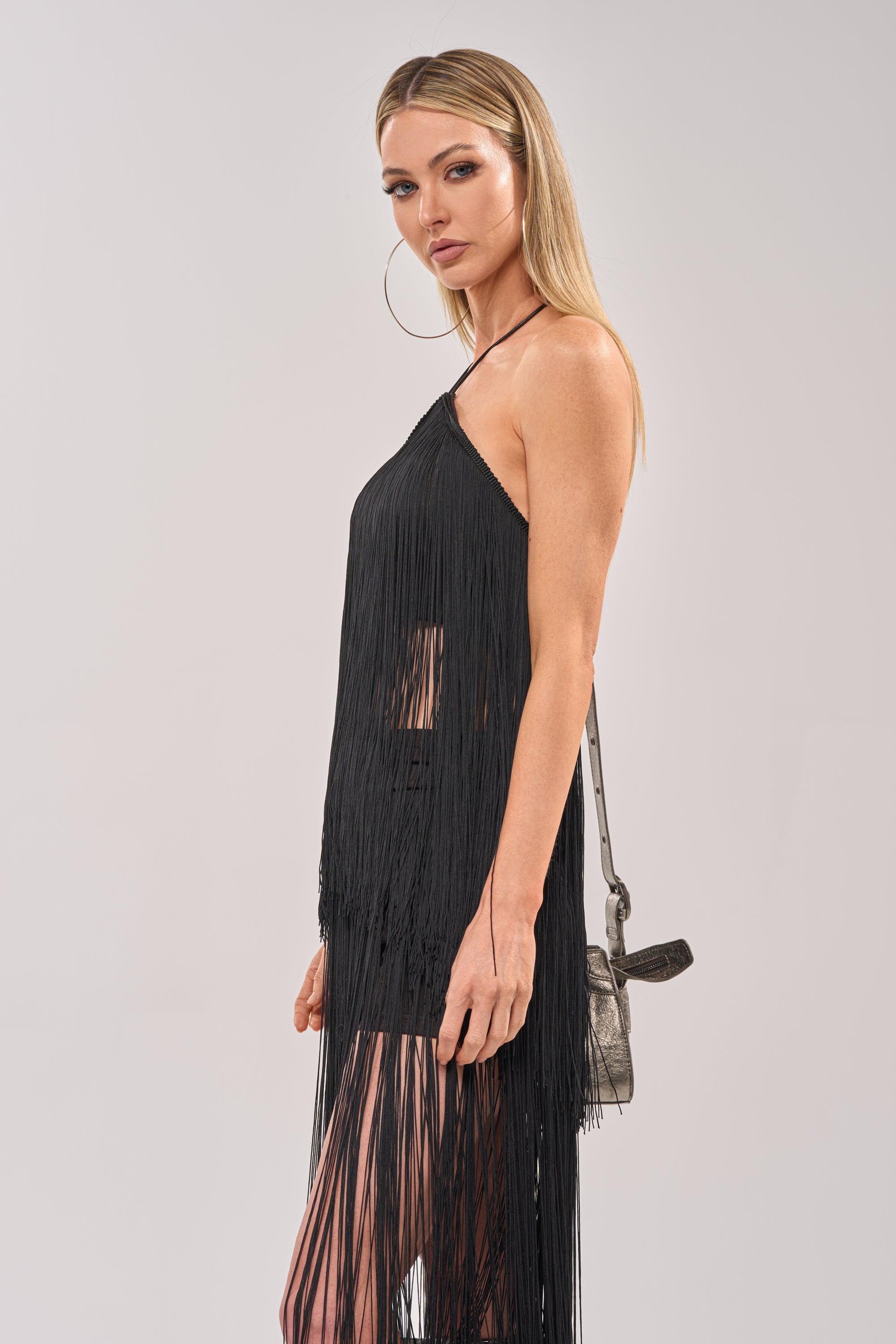A woman with long blonde hair wears the ON THE EDGE FRINGE HALTER TOP IN BLACK, paired with large hoop earrings and a small metallic purse, standing against a light gray background. 3