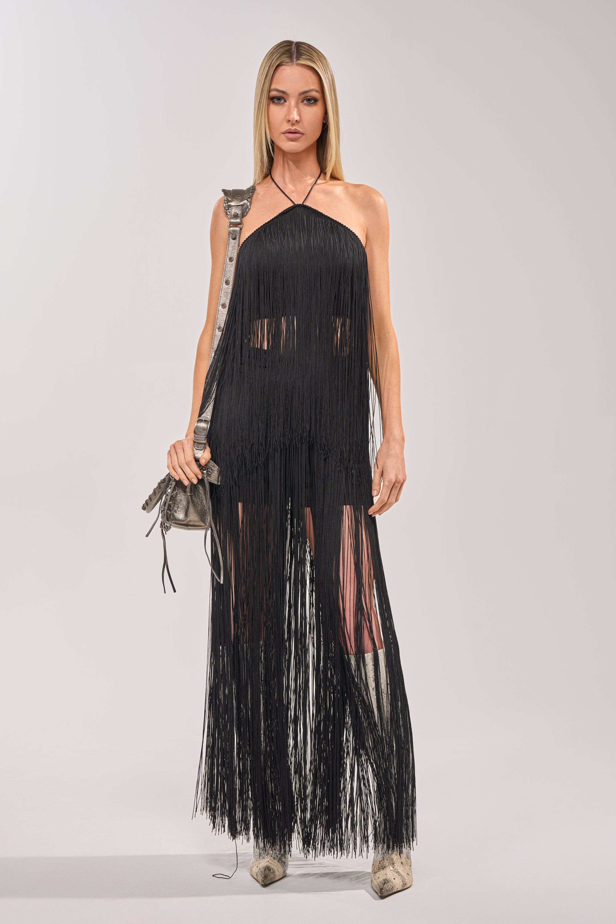 A woman faces forward in the ON THE EDGE FRINGE HALTER TOP IN BLACK, styled with a silver bag and light boots. Her occasion wear look highlights the statement top against a plain, light background. 5