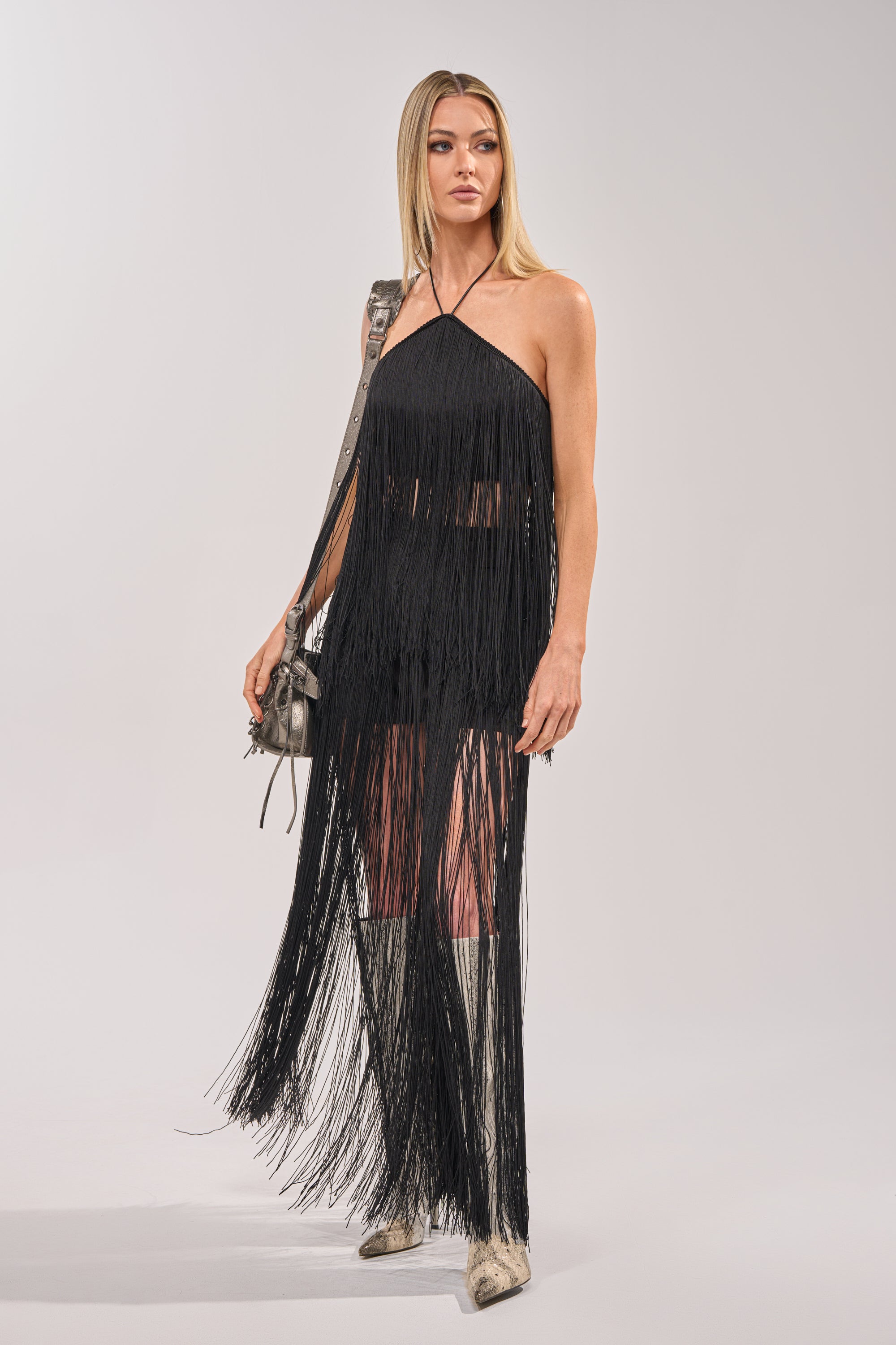 A woman in the ON THE EDGE FRINGE HALTER TOP IN BLACK, paired with silver boots and a small silver bag, stands against a plain light background. Her loose straight blonde hair completes this chic occasion look with subtle statement styling. 6