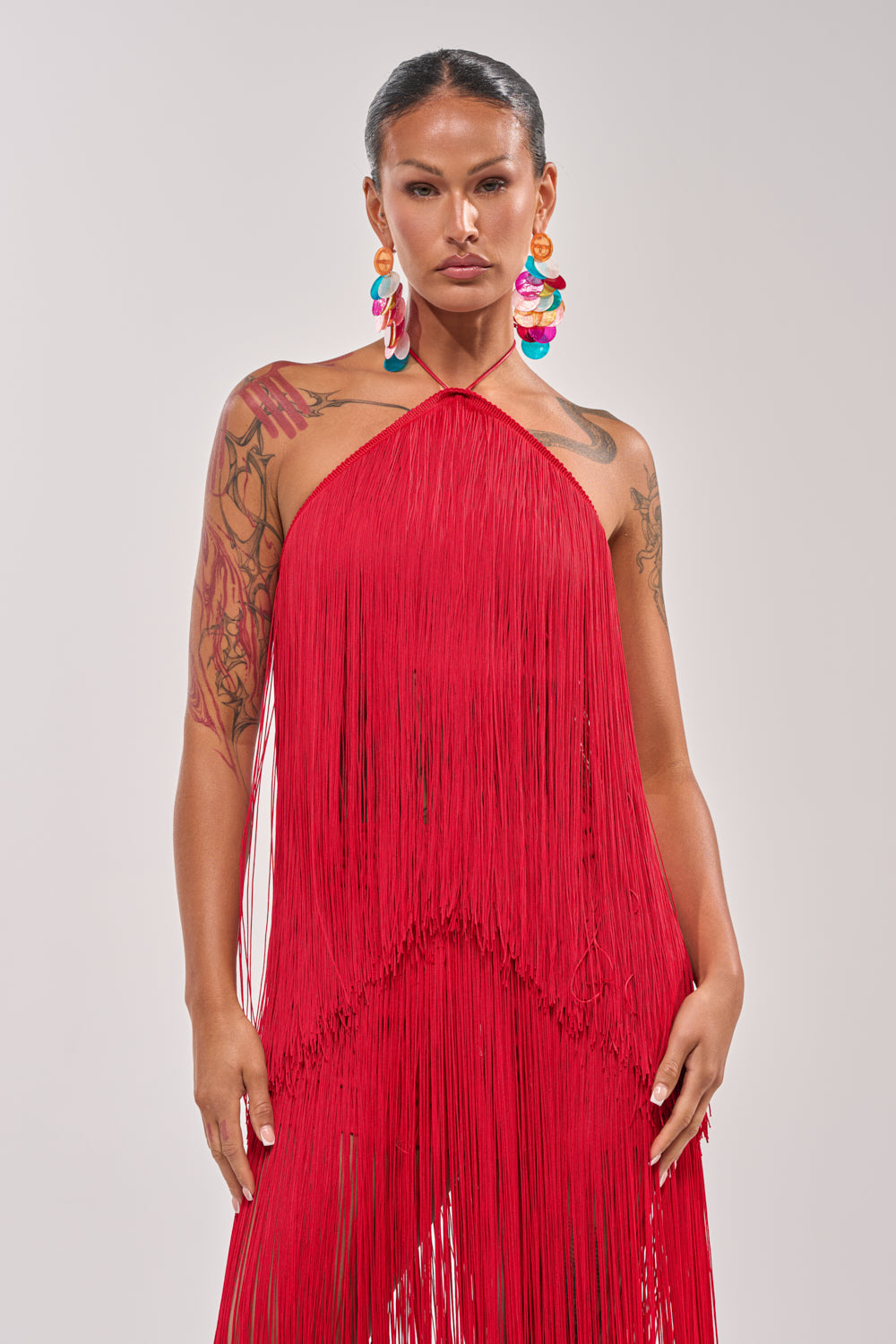 A tattooed woman with slicked-back hair wears the ON THE EDGE FRINGE HALTER TOP in red and colorful statement earrings, standing against a plain light background—perfect for festival looks or bold statement outfits. 1