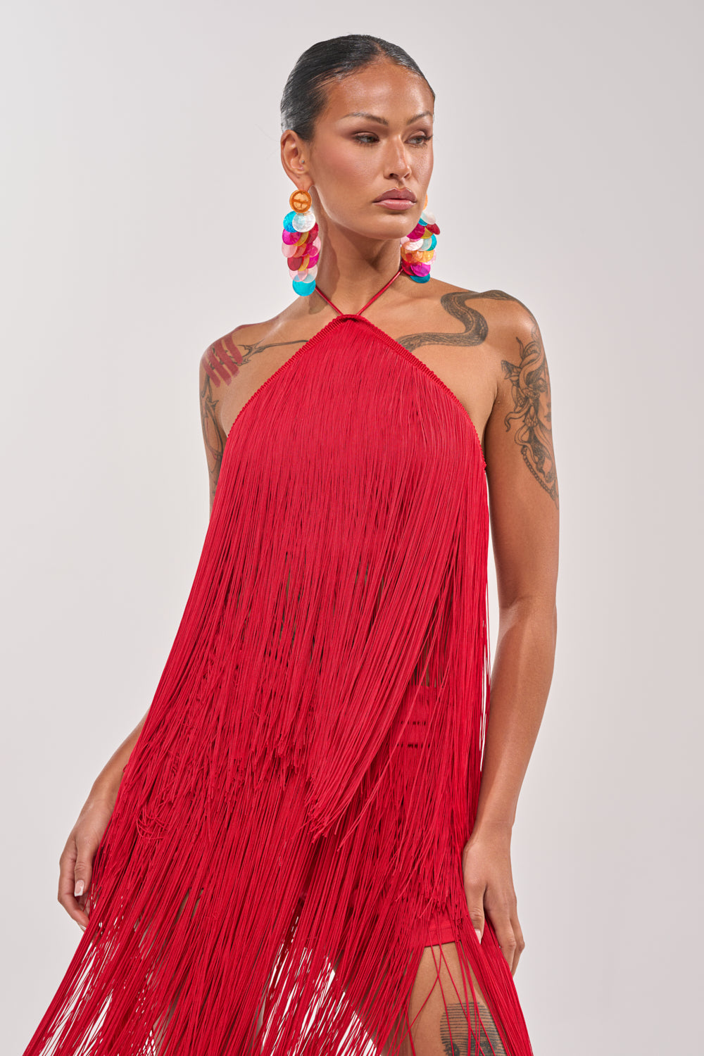 A tattooed woman wears the ON THE EDGE FRINGE HALTER TOP in red with bold, colorful hoop earrings, creating a statement look against a plain light background. 2