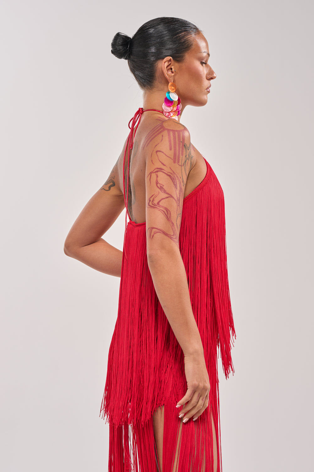 A woman with dark hair in a bun wears the ON THE EDGE FRINGE HALTER TOP and colorful earrings. Tattoos cover her arm as she stands in profile against a light background, highlighting bold festival style. 3