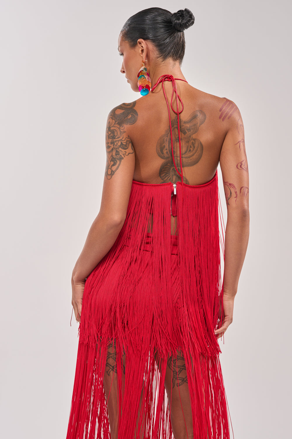 A woman with tattoos wears the ON THE EDGE FRINGE HALTER TOP and colorful earrings, her hair in a neat bun, standing with her back to the camera against a light gray background. 4