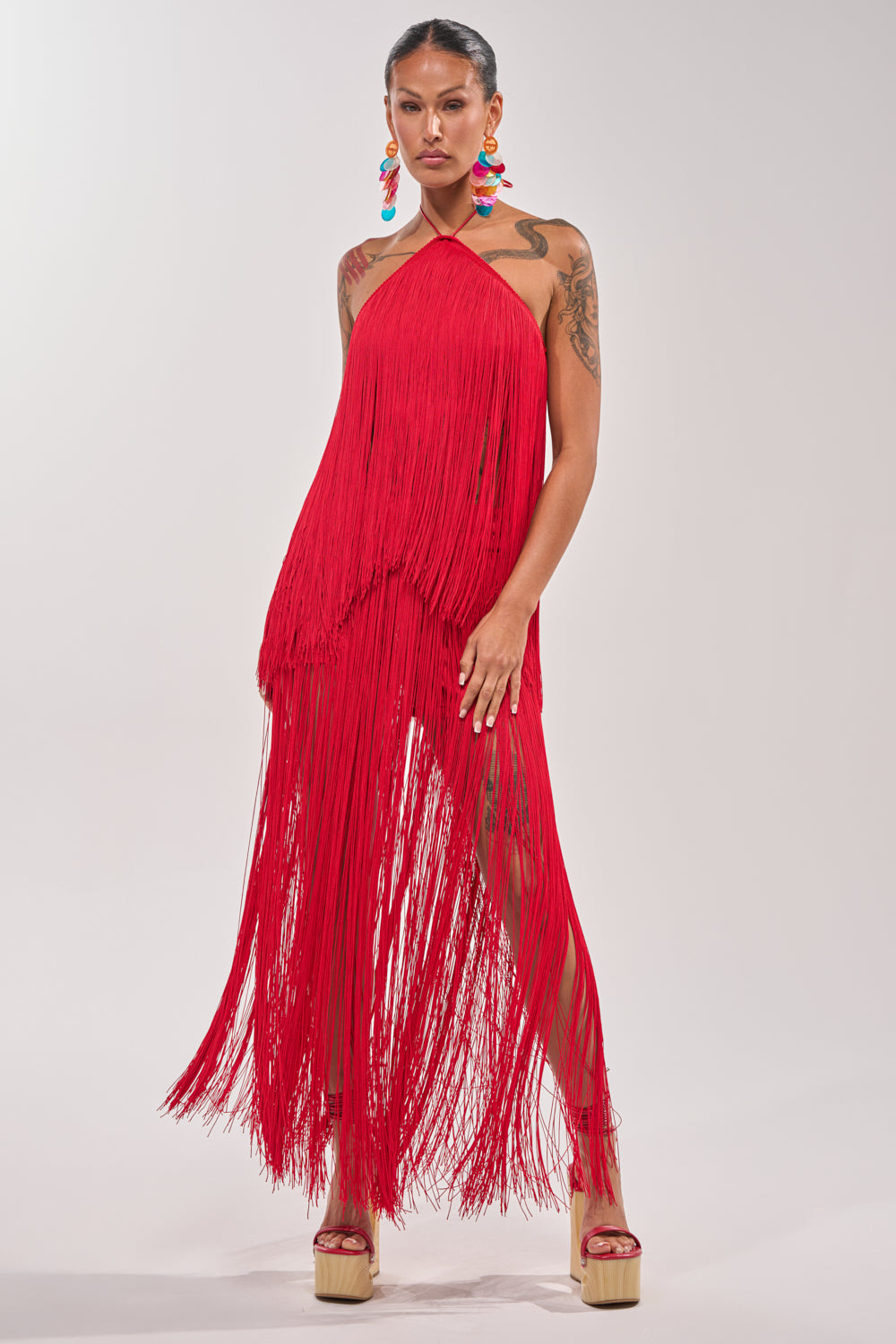 Against a plain backdrop, a woman wears the ON THE EDGE FRINGE HALTER TOP in bright red with a matching skirt, platform heels, and colorful statement earrings. Her arm tattoos add edge to this bold, confident festival look. 5