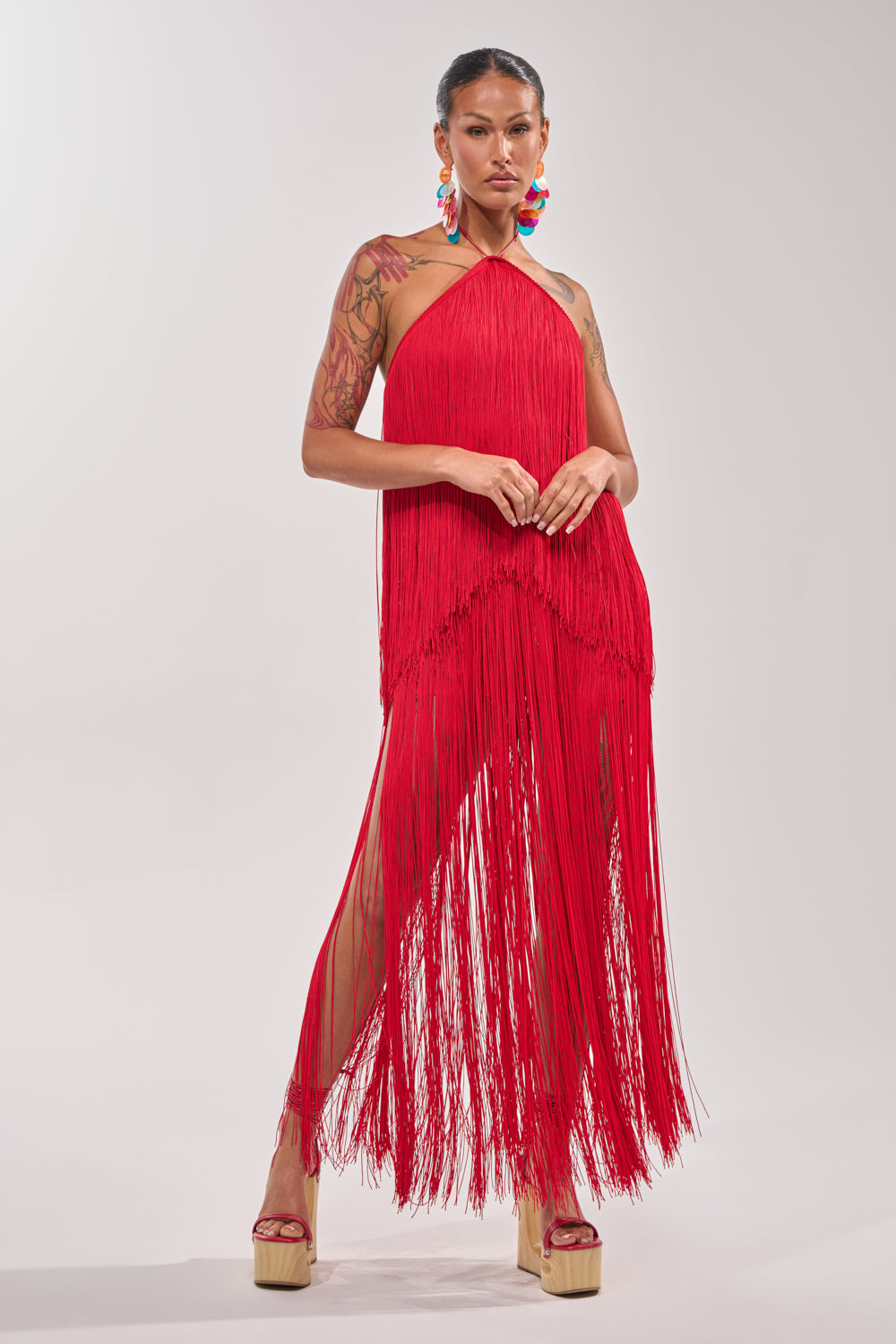 A person stands confidently in the ON THE EDGE FRINGE HALTER TOP and matching platform heels, bold earrings, and visible arm tattoos against a plain light background—perfect for standout festival outfits. 6