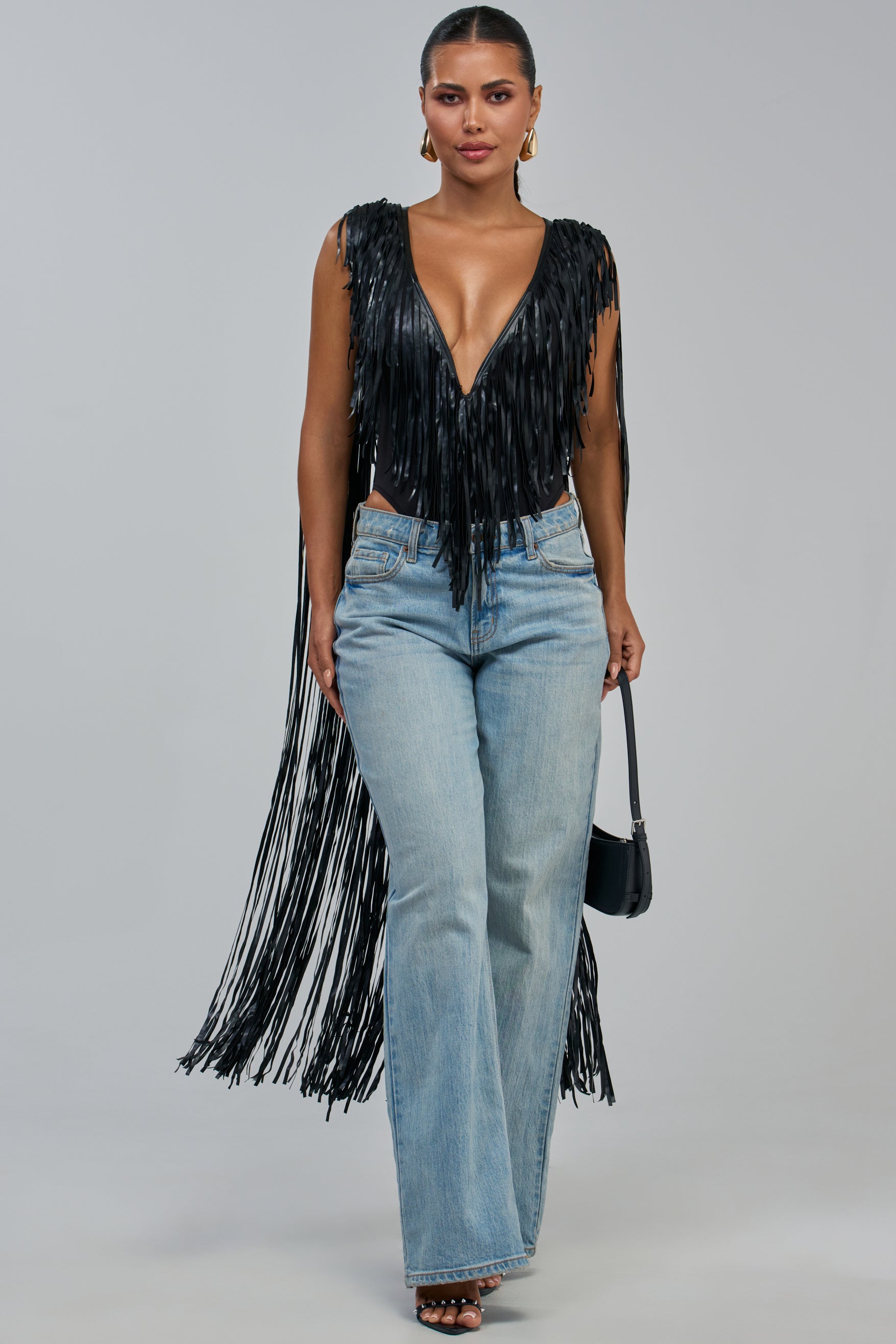 A woman poses in light blue jeans and the MEET ME IN THE DESERT PU FRINGE BODYSUIT, a black top featuring long fringe details for a festival-inspired look. She accessorizes with a small black handbag, black open-toe heels, gold earrings, and has her hair pulled back. The background is plain light gray.