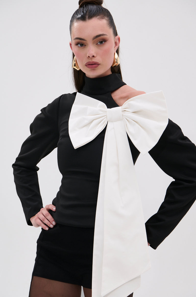 A woman wears the DAYANNE MOCK NECK BOW TOP, a black long-sleeve mock neck with a striking white bow and sash draped over one shoulder, styled with black shorts, sheer tights, gold earrings, and her dark hair pulled back for an elegant look. 0