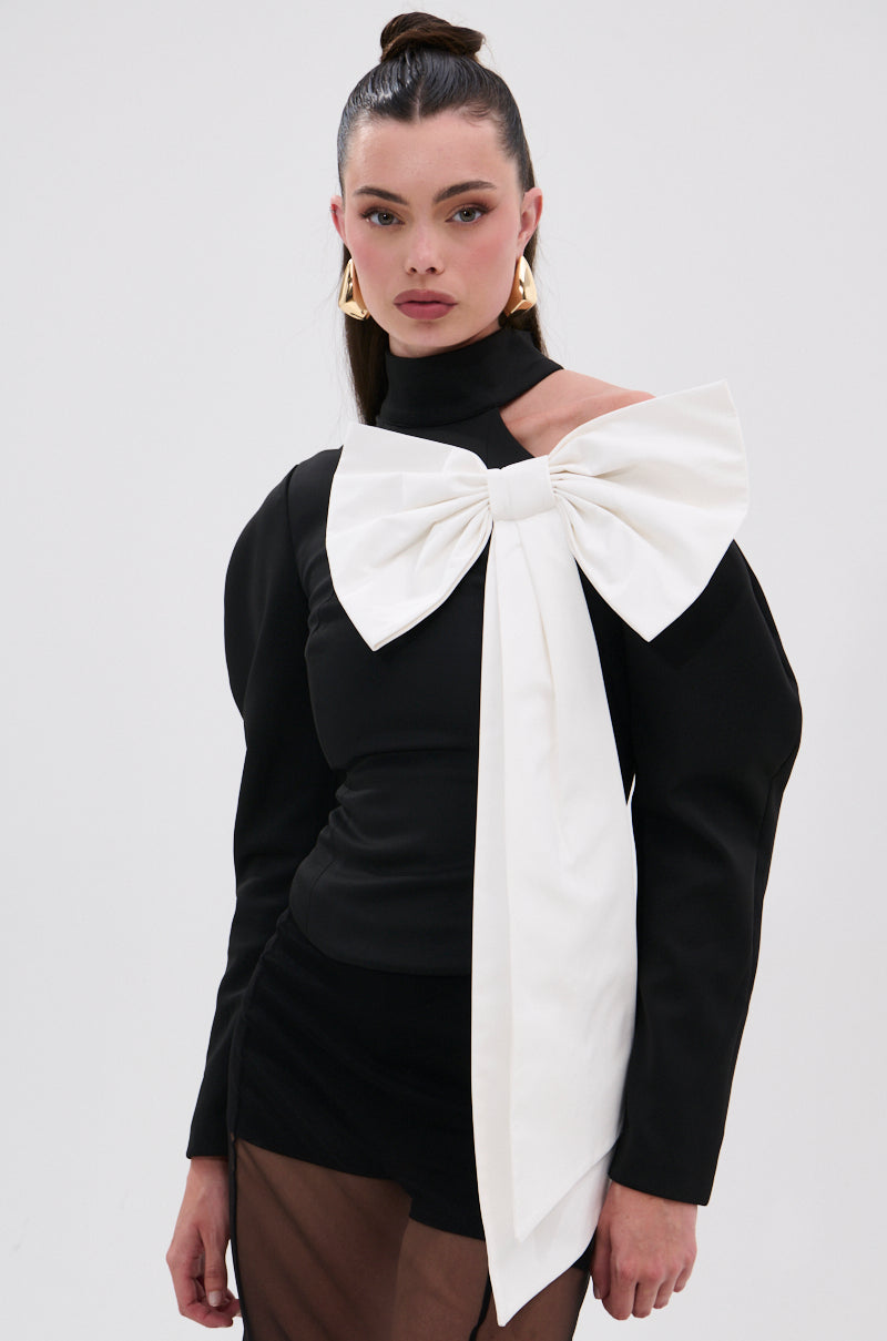 A woman models the DAYANNE MOCK NECK BOW TOP—black, long-sleeved with a large white shoulder bow—paired with gold earrings and a sheer black skirt. Her hair is styled in a high bun against a plain white background. 1