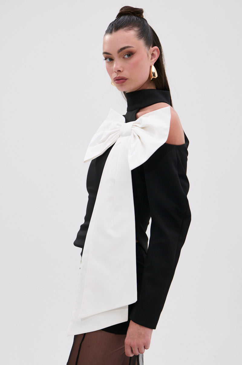 A woman with long dark hair in a high ponytail wears the DAYANNE MOCK NECK BOW TOP, featuring sheer sleeves and a large white bow across the shoulder. She stands against a plain white background with a neutral expression, styled for eveningwear. 2