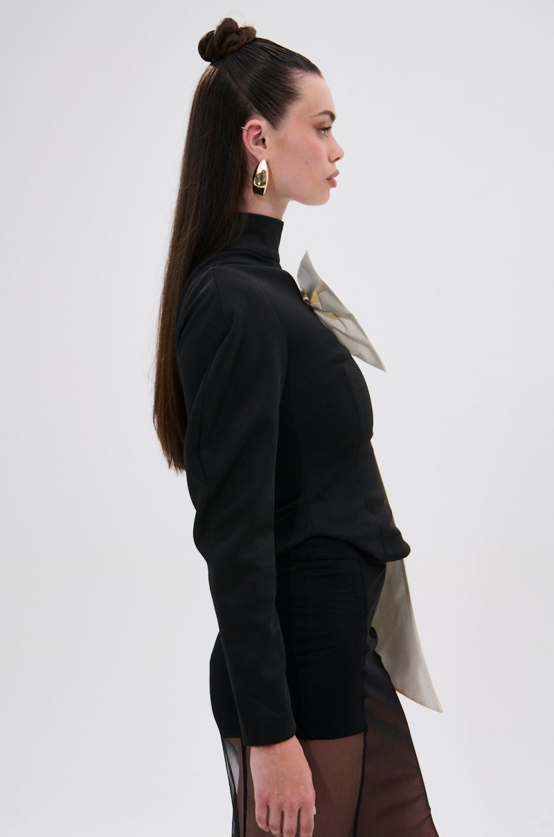 A woman stands in profile against a plain background, wearing the DAYANNE MOCK NECK BOW TOP—an elegant black piece with sheer panels, an asymmetrical cut, and a large metallic bow at the neck. Her long dark hair is half-up with bold gold earrings. 3