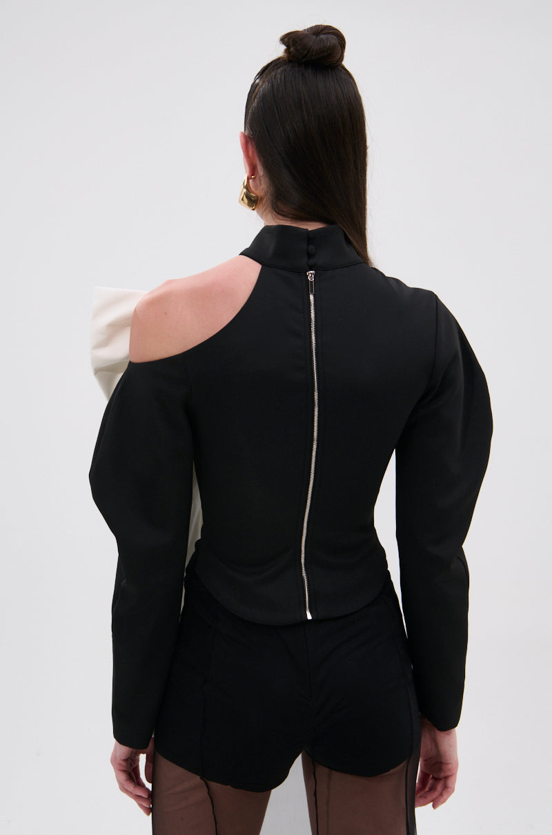 A person with long dark hair in a topknot is seen from behind wearing the DAYANNE MOCK NECK BOW TOP, a black asymmetric eveningwear piece with a mock neck, back zipper, cut-out shoulder detail, paired with black shorts and sheer tights. 4