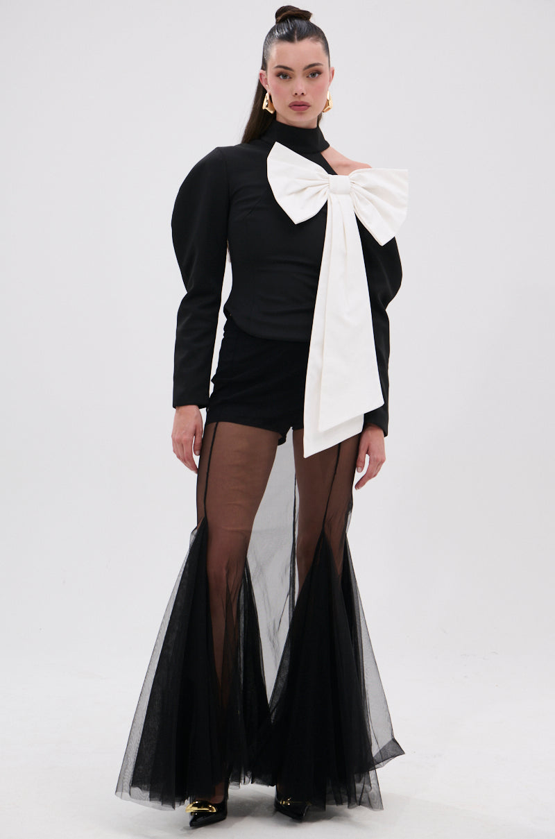 A woman wears the DAYANNE MOCK NECK BOW TOP—black with puffed sleeves and a large white bow—paired with sheer black flared pants over black shorts. She has slicked-back hair and gold earrings, posing against a plain white background. 5