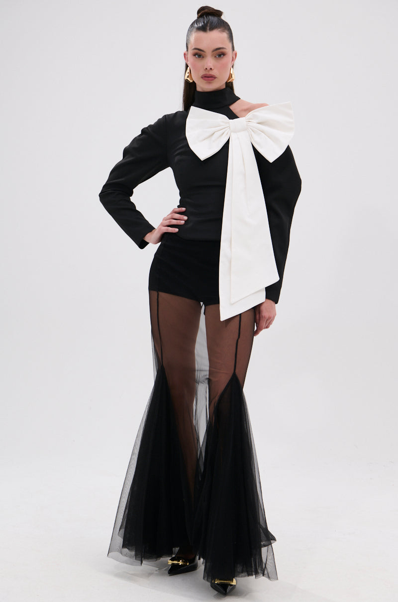 A woman models the DAYANNE MOCK NECK BOW TOP—an elegant black top with a statement white bow—paired with sheer flared pants, gold pointed heels, and gold earrings. Her sleek hair and confident pose complete the look against a plain white background. 6