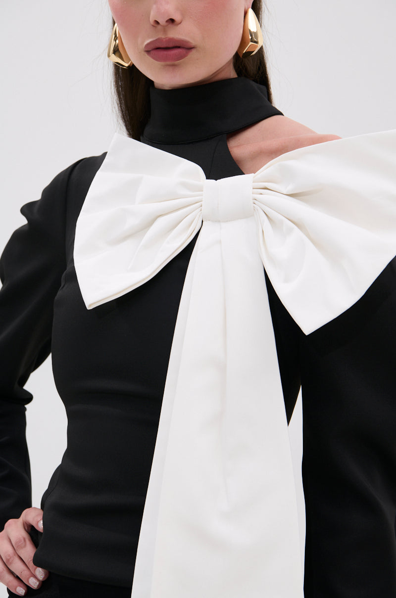 A woman models the DAYANNE MOCK NECK BOW TOP—black with a striking white bow on the shoulder—paired with gold geometric earrings and neutral lipstick, giving her an elegant evening look against a soft, light background. 7