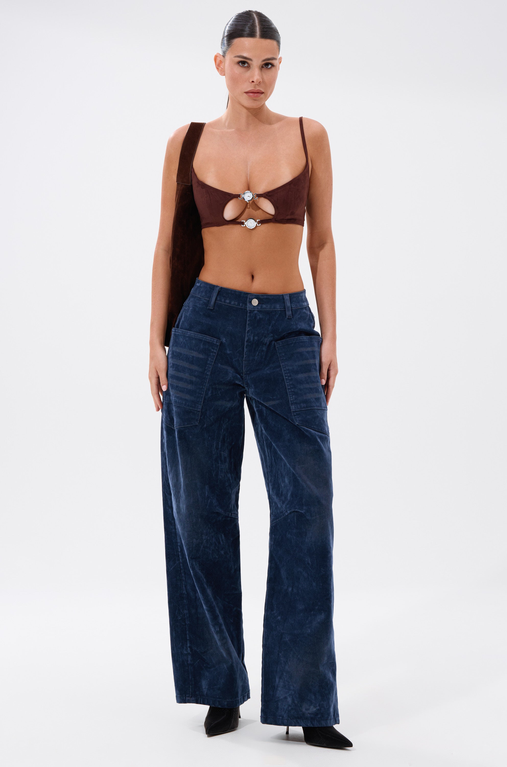 A woman with sleek pulled-back hair and a serious expression wears the READ BETWEEN THE LINES CLOCK BRALETTE, high-waisted wide-leg blue pants, black pointed shoes, and a brown shoulder bag against a plain white background. 6